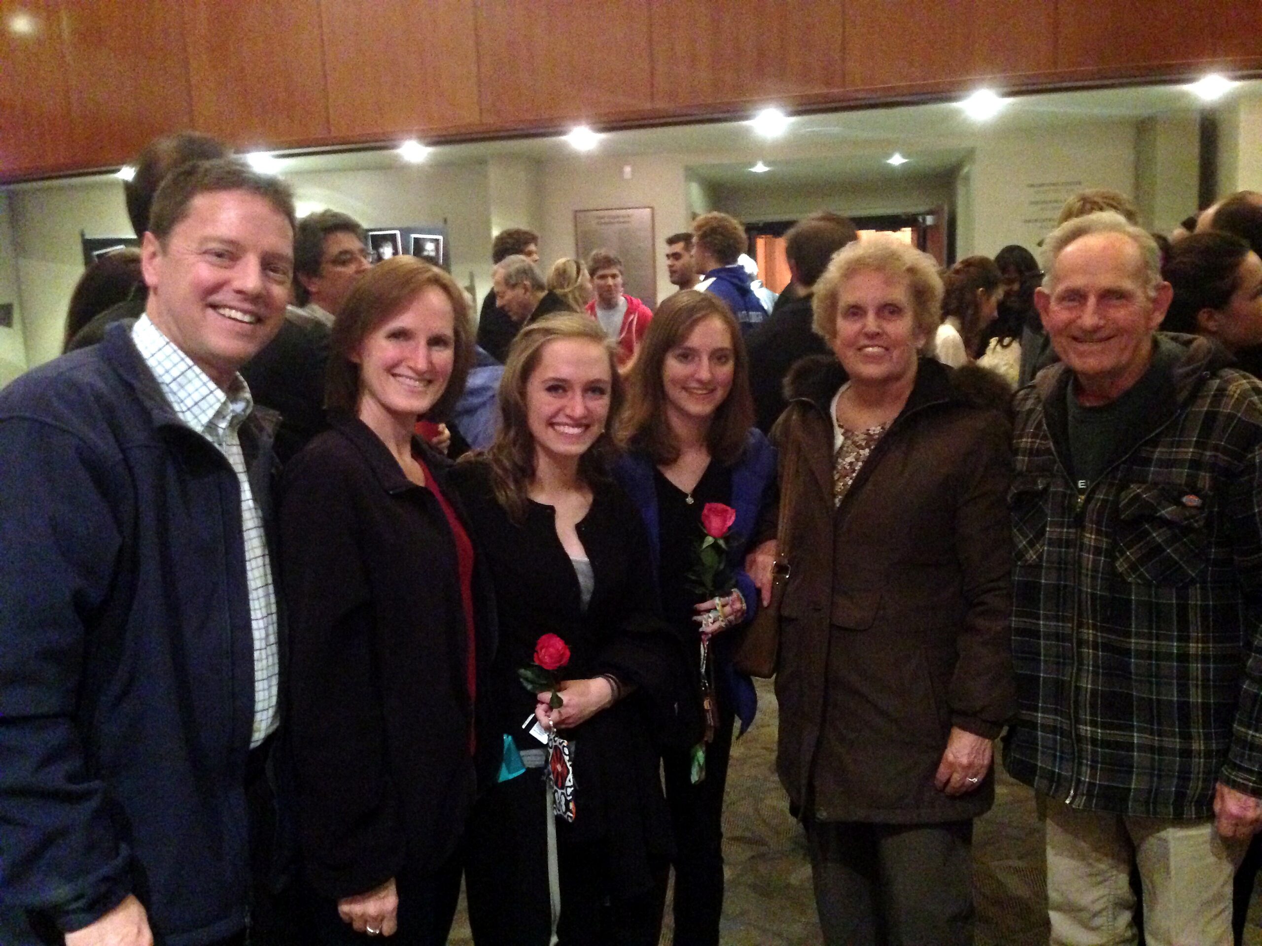Group of six people smiling for a photo in a busy indoor lobby; two women hold red roses.