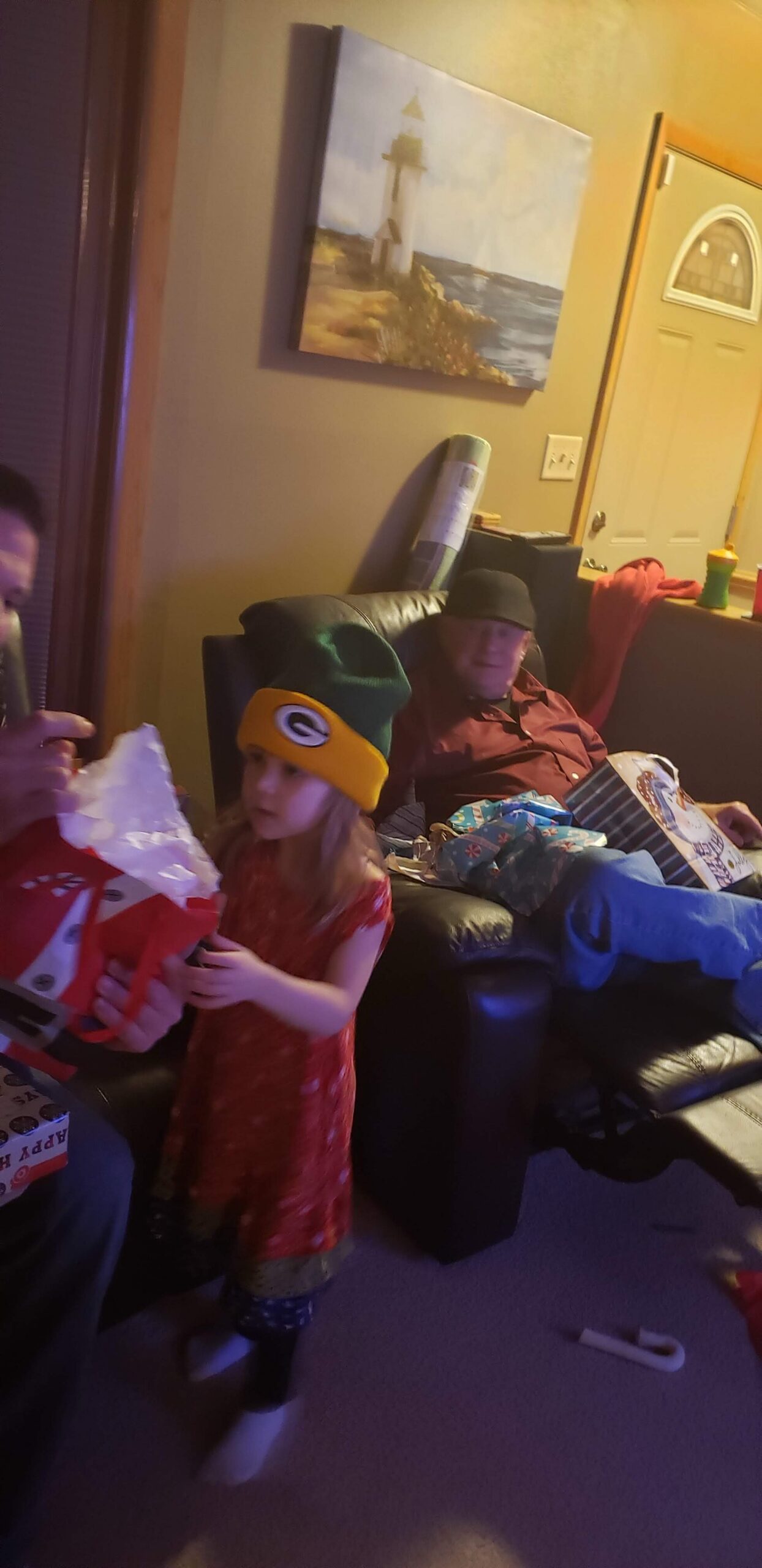 Young girl in a green and yellow hat opens a red gift bag on a couch, as an older man sits nearby amid scattered presents in a living room.