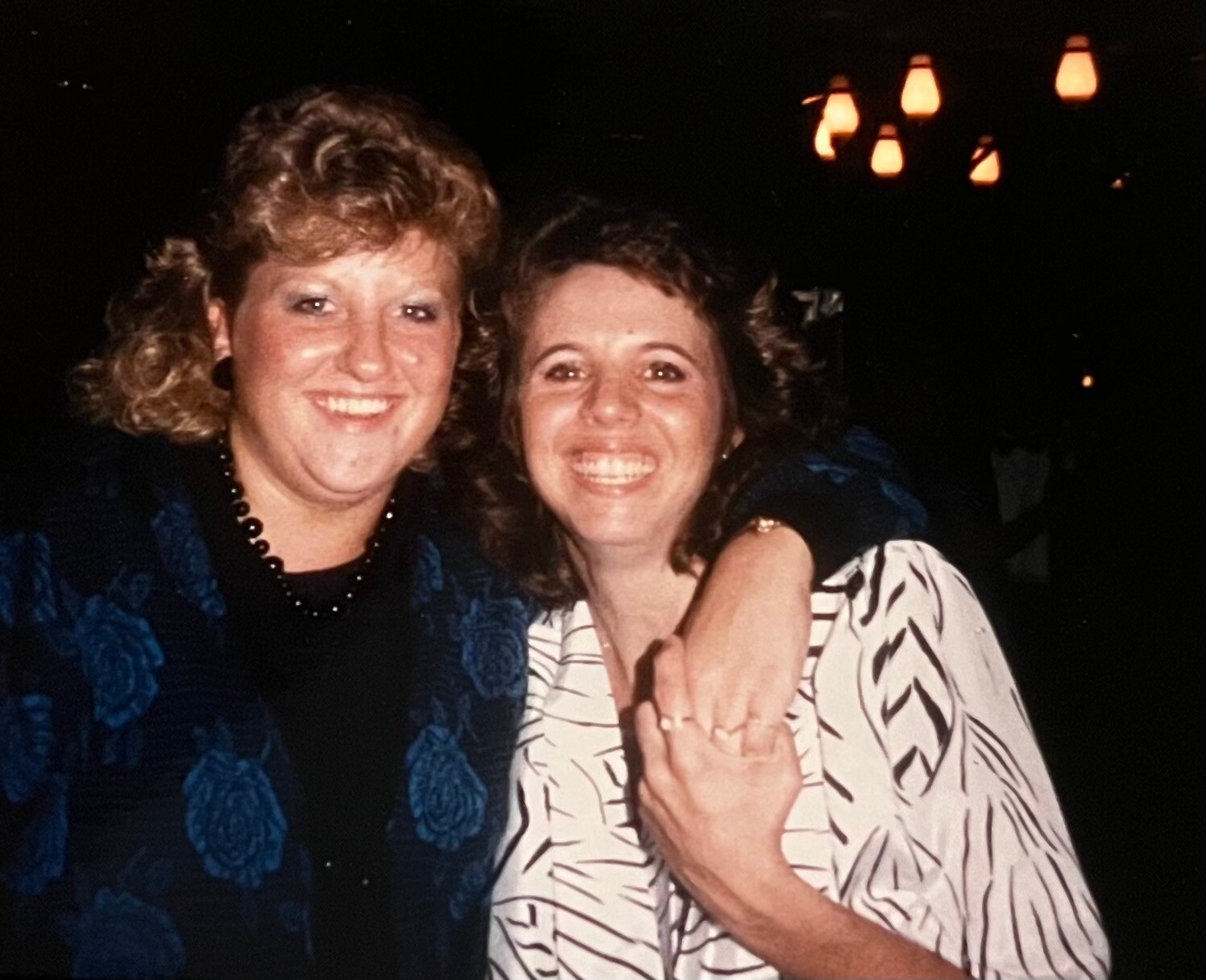 Two women smiling and embracing at a dimly lit social event, one in a blue floral jacket and the other in a white striped blouse, with hanging lanterns in the background.