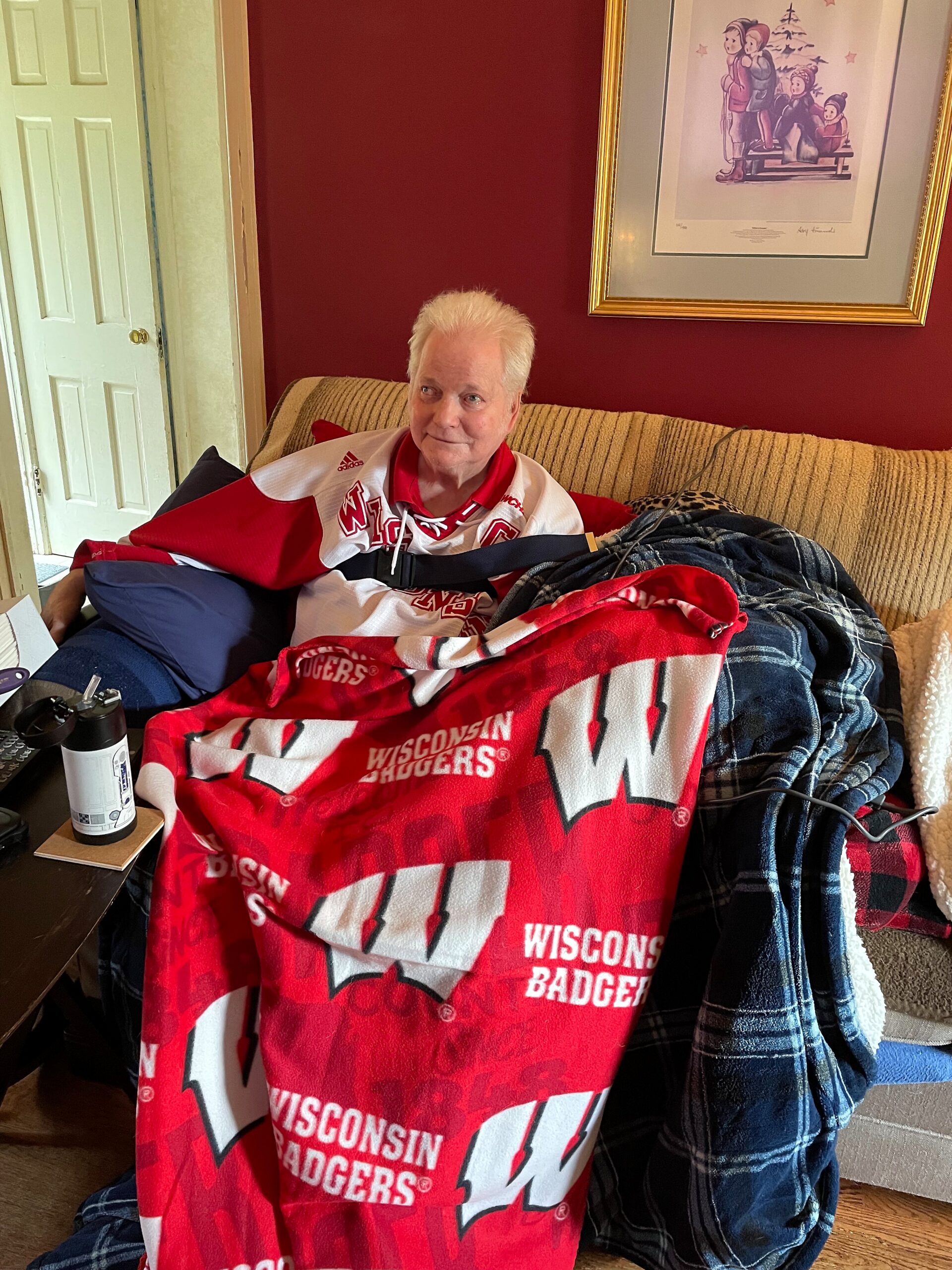 Elderly man with white hair sitting on a beige sofa, wrapped in a red Wisconsin Badgers blanket in a living room.