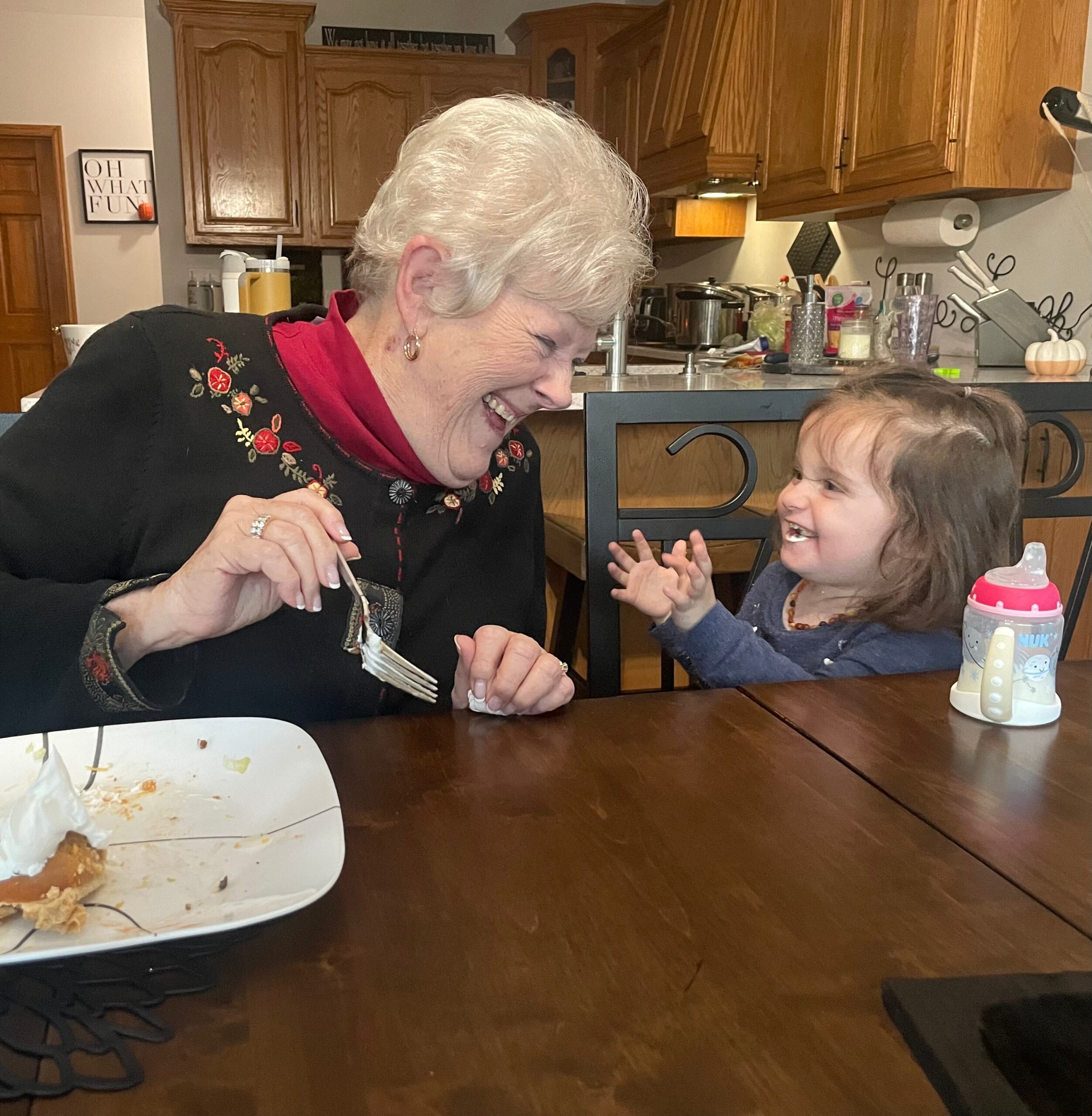 Grandmother and young child smiling at a kitchen table with a cake plate between them on a dark wood table.