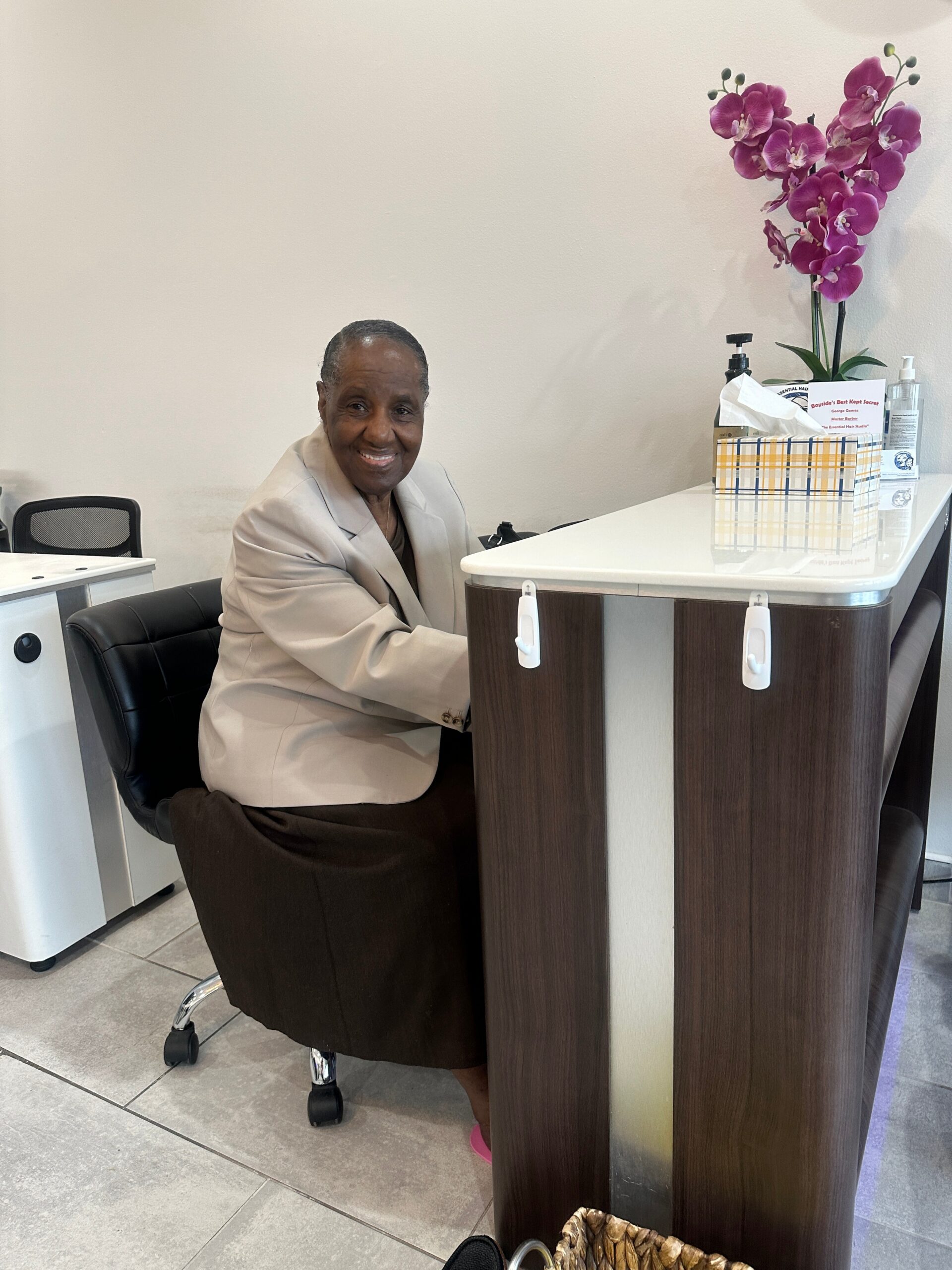 Smiling older woman in a beige blazer sits at a reception desk with pink orchid decor on the counter.