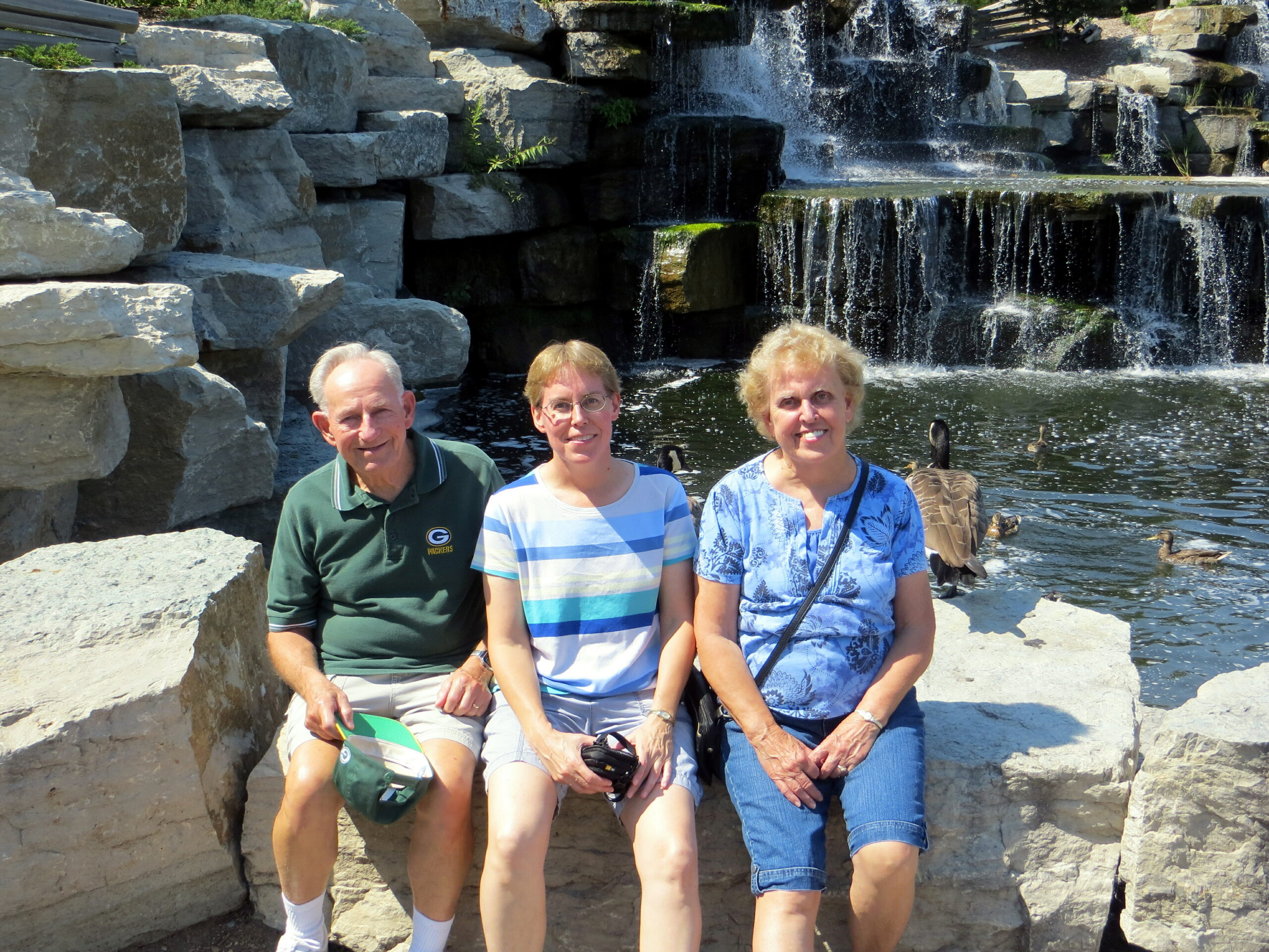 Three adults posing on a rocky ledge beside a multi-tiered waterfall in a park setting.
