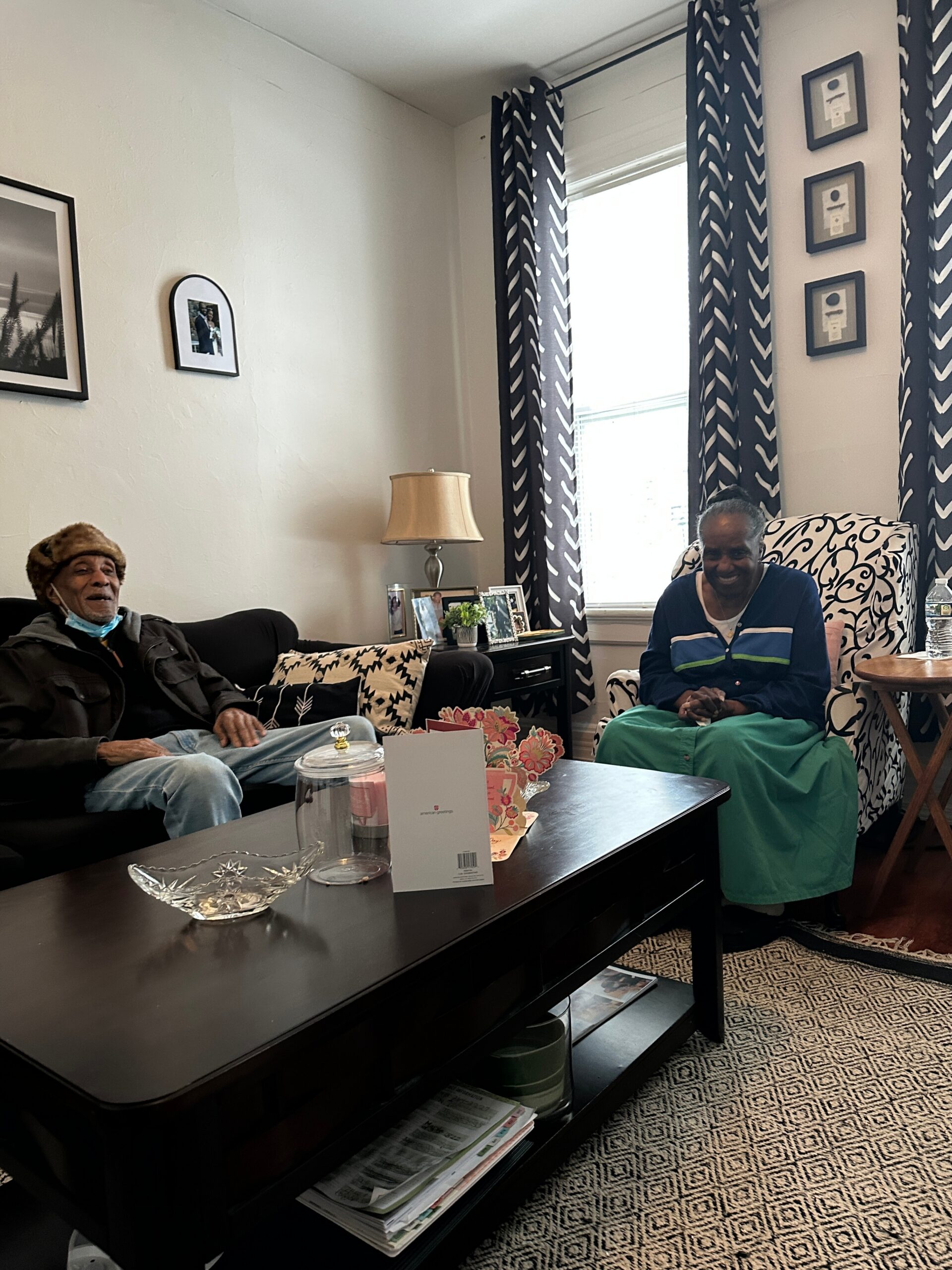 Two elderly people sit in a cozy living room—a man on a sofa left and a woman in a patterned chair on the right, smiling.