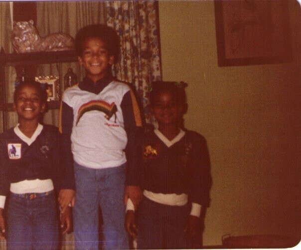 Three children standing indoors, smiling for the camera; two wear dark uniforms with white belts, center child in a light shirt with a rainbow graphic.