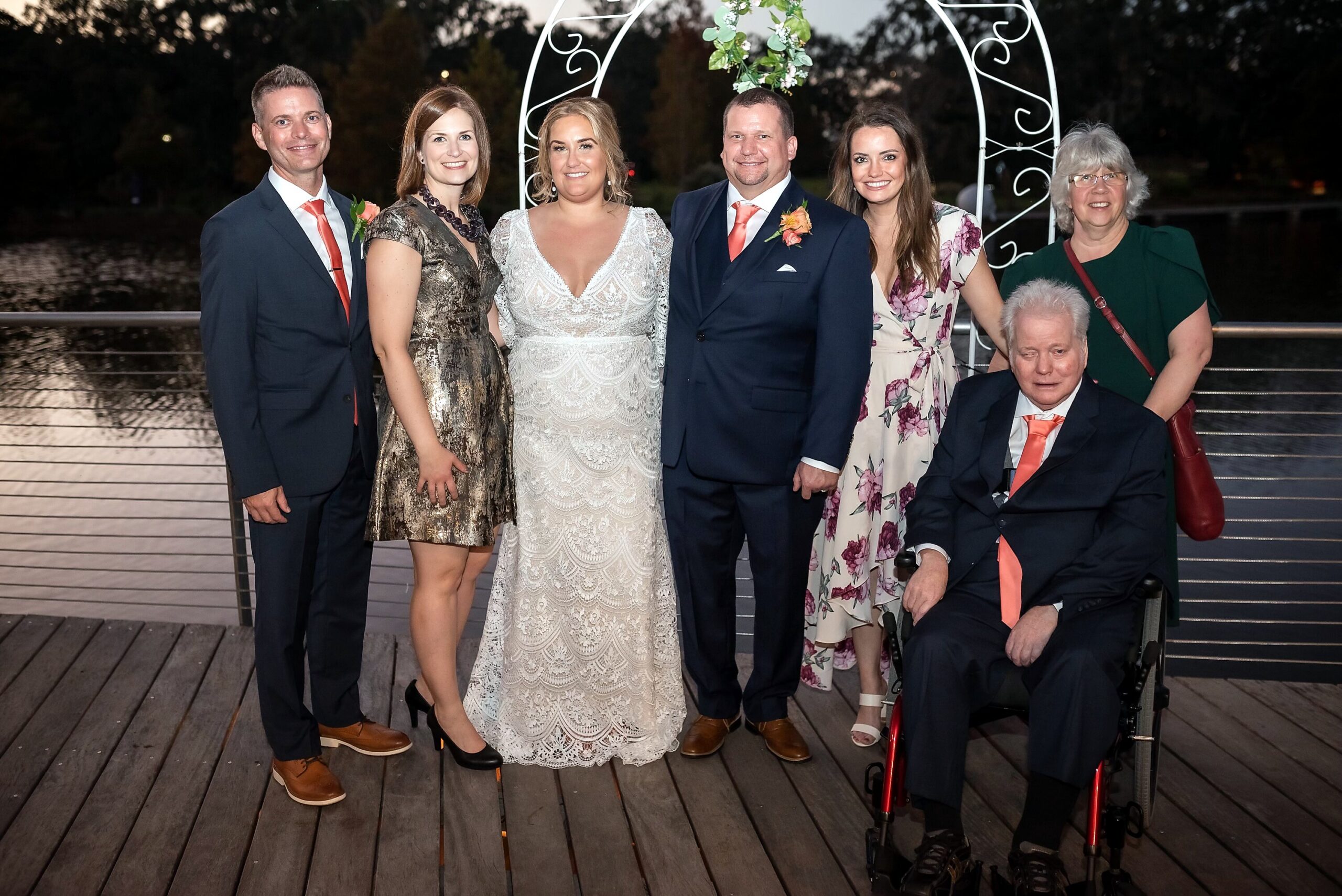 Group of six adults posing on a wooden boardwalk at night, two in formal dresses and three men in suits, plus an older man in a wheelchair on the right.