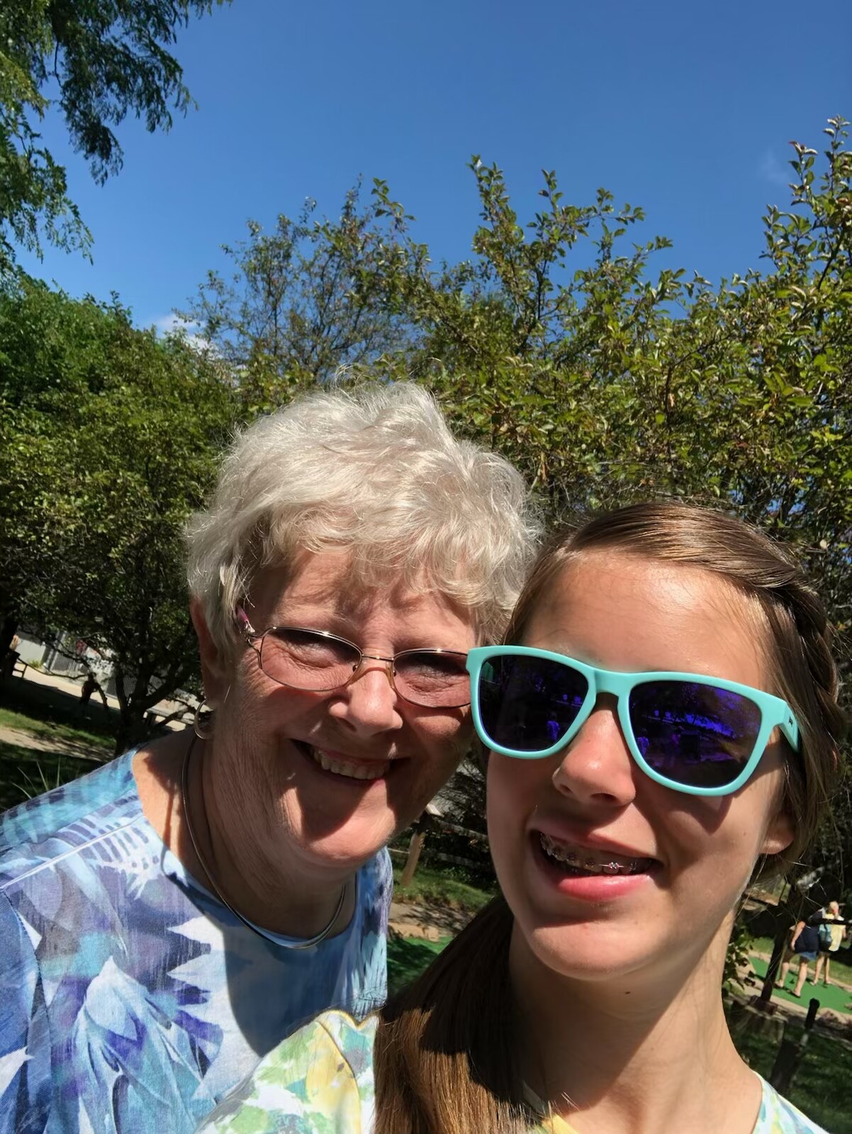 Smiling older woman and younger girl taking a sunny outdoor selfie with green trees and blue sky behind them.