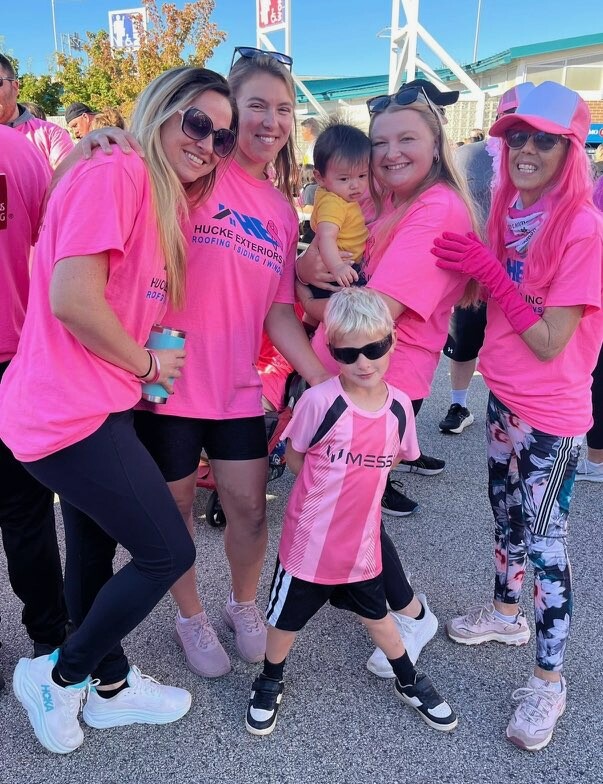 Group of smiling volunteers in pink shirts posing with a small child at an outdoor event.