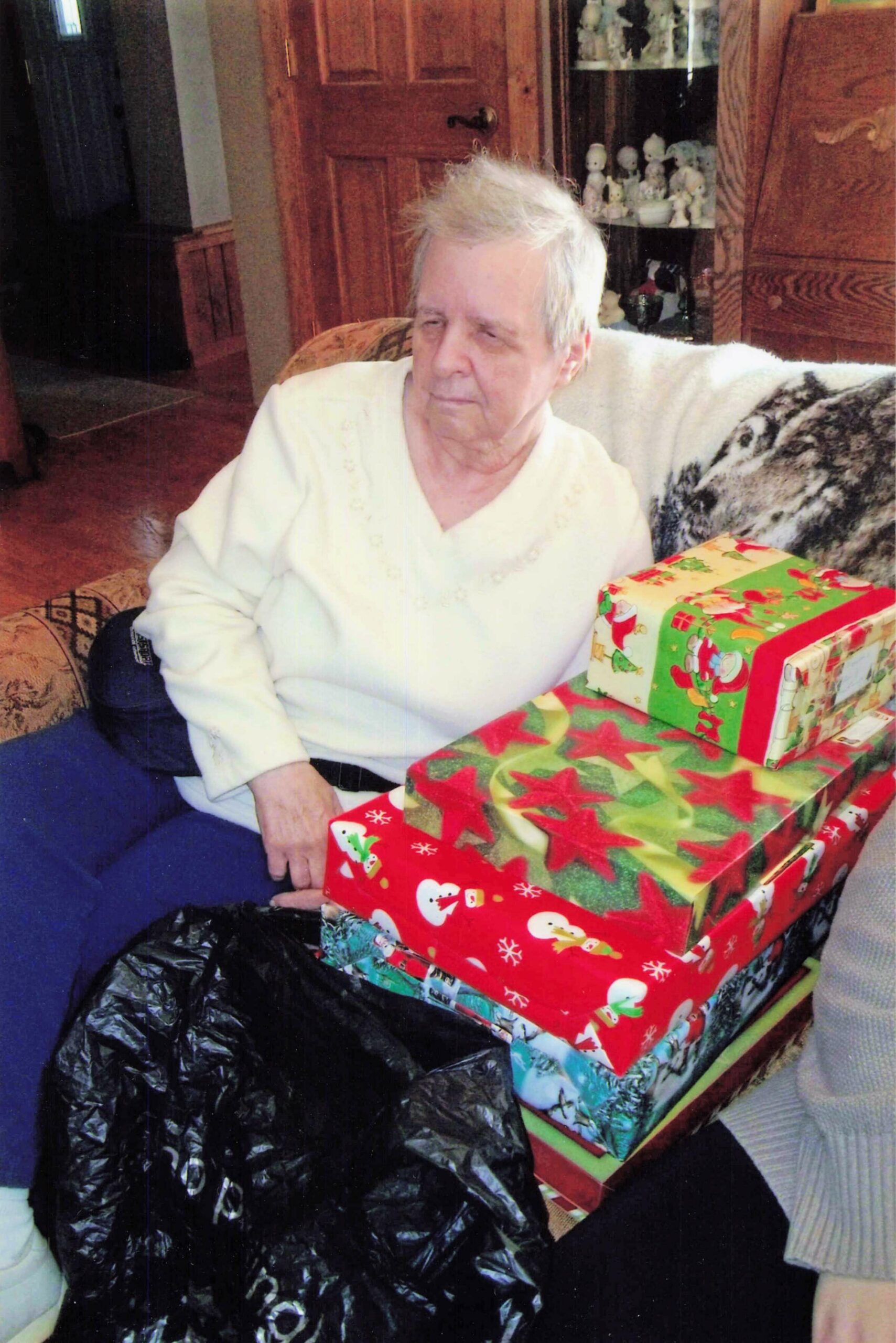 Older person seated on a sofa with wrapped Christmas gifts nearby in a cozy living room.