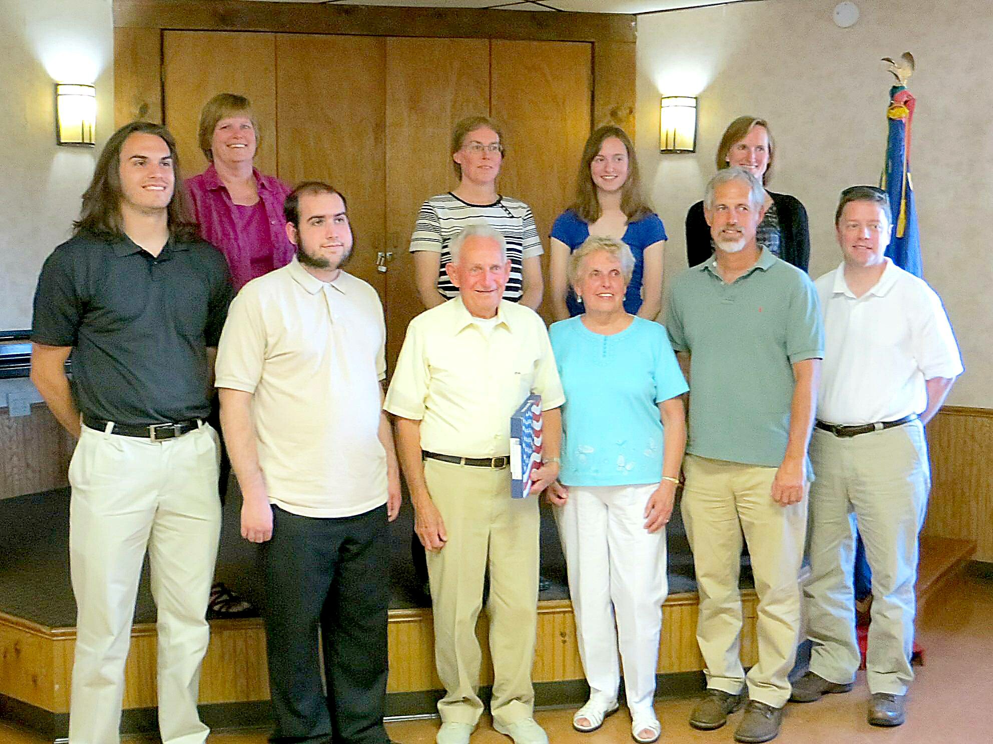 Group of adults posing for a photo in an indoor room with wood paneling and a flag in the corner, all smiling.