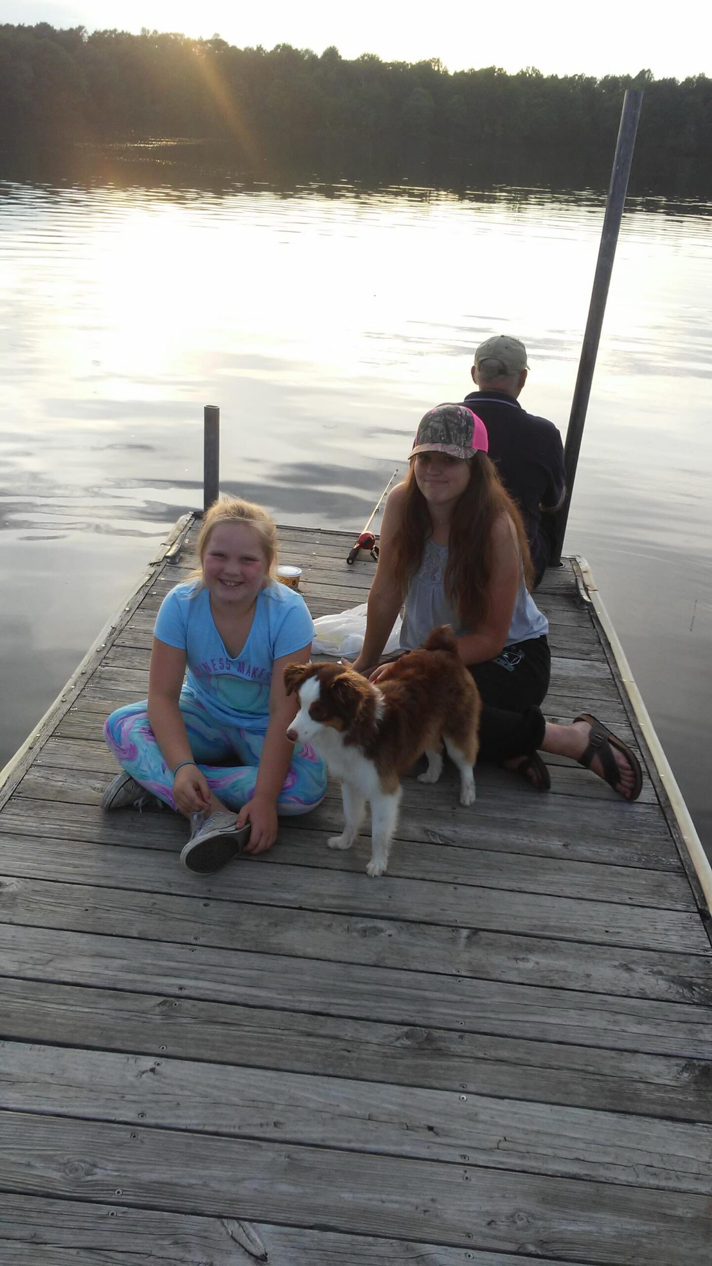 Two smiling girls sit on a wooden dock by a calm lake with a brown-and-white puppy nearby; a man sits fishing in the background as sunlight reflects on the water.