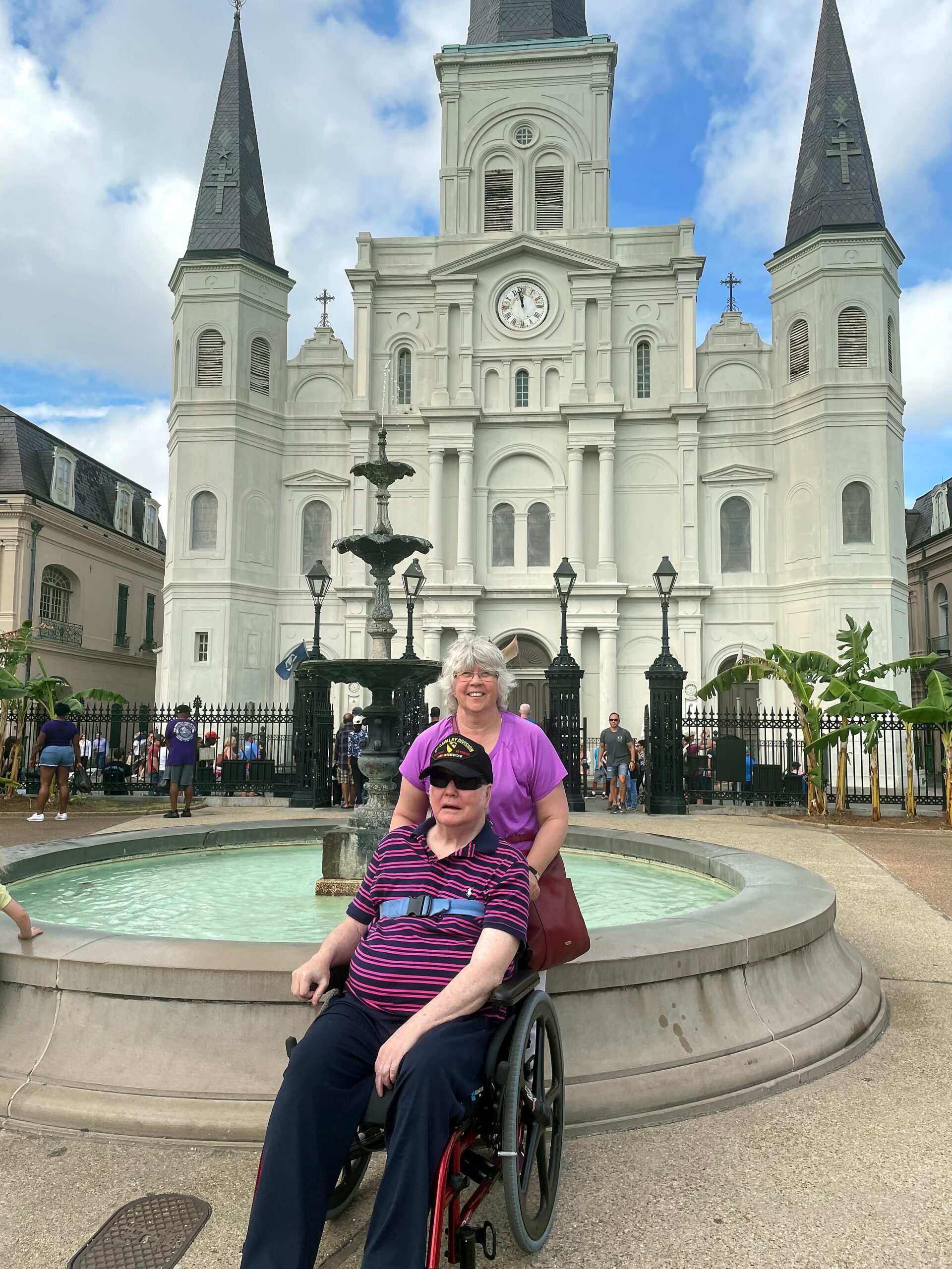 White church with two tall spires and a central clock, in a plaza with a fountain; two women in wheelchairs smiling at the camera.