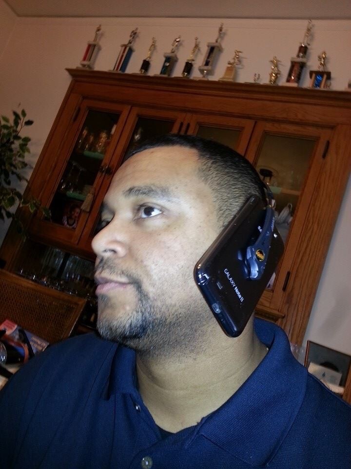 Man in a navy polo speaks on a large black smartphone clipped to his ear at home, with a wooden cabinet and trophies in the background.