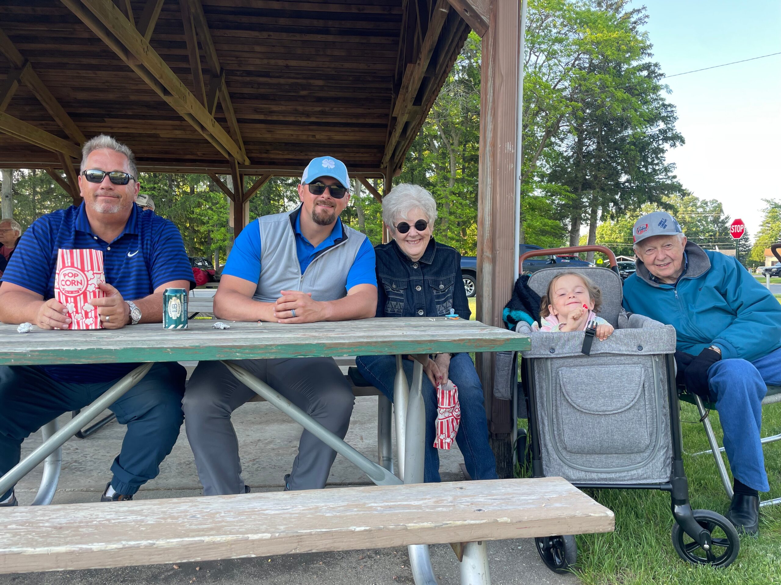 Group of five people at a wooden picnic table under a pavilion, smiling at the camera with snacks and drinks on the table behind them.