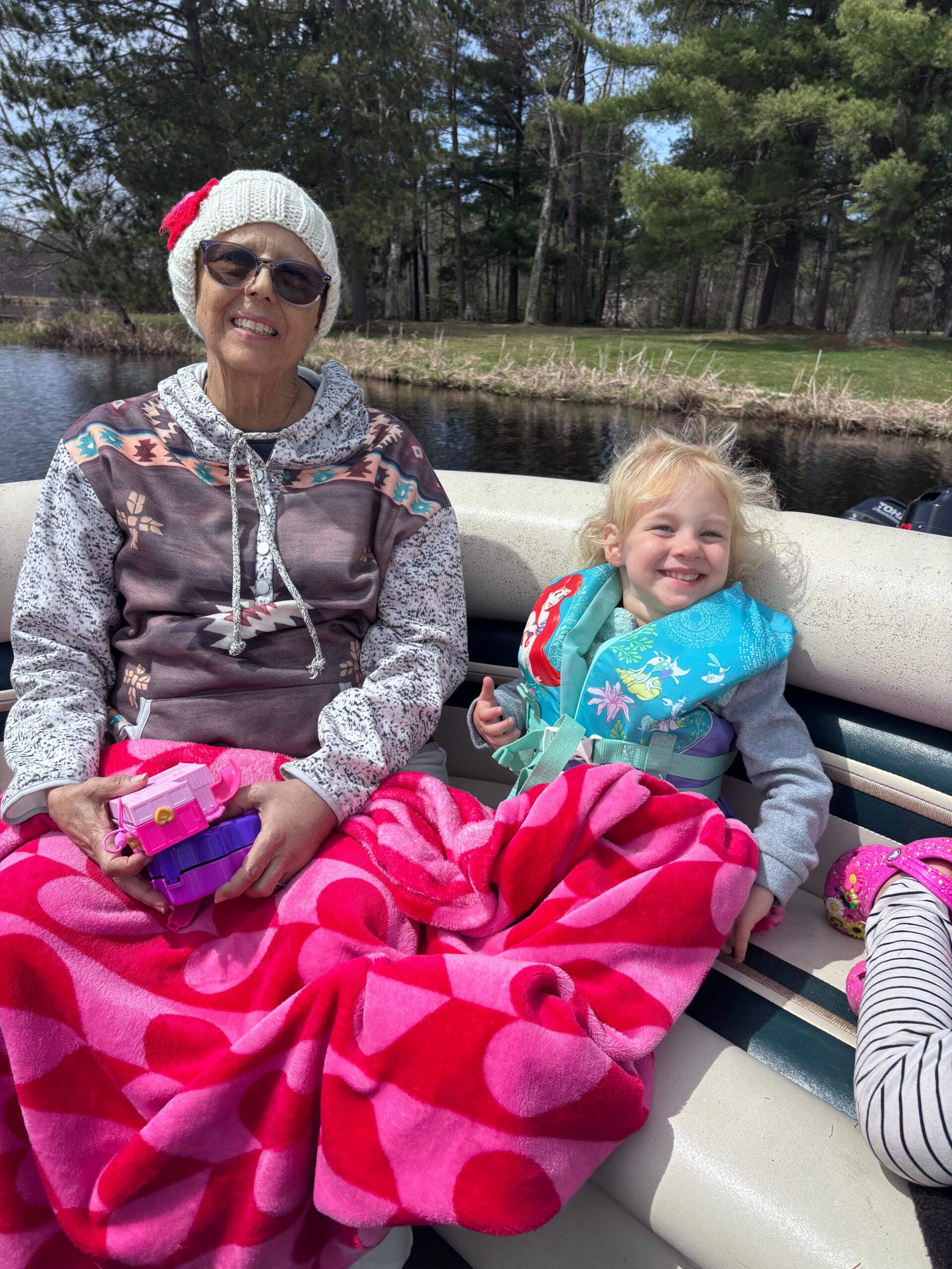 Elderly woman in a white knit hat and sunglasses sits on a boat beside a smiling child in a life jacket, wrapped in a pink blanket.