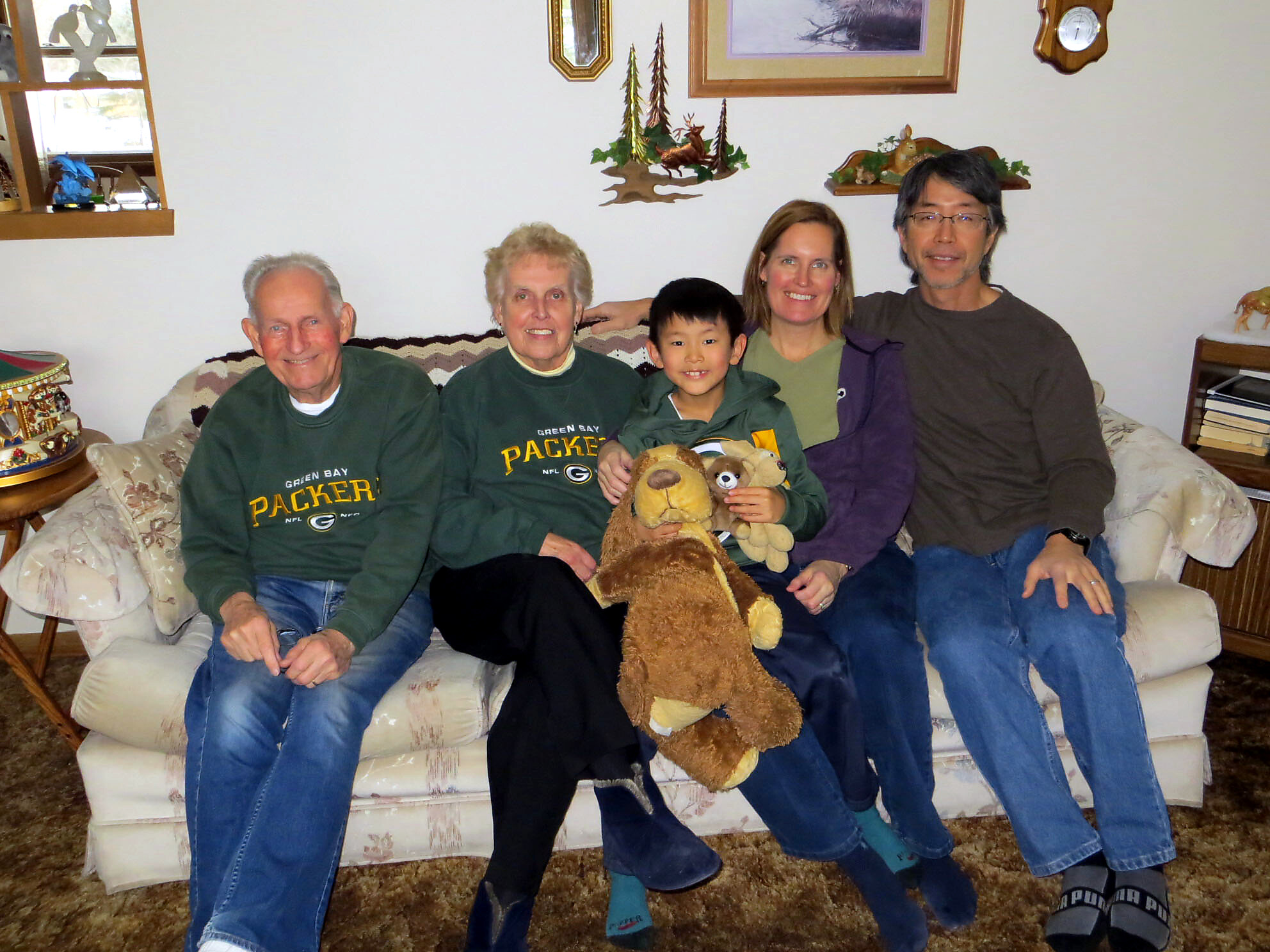 Five family members sit closely on a cream sofa, smiling, holding stuffed animals in a cozy living room.