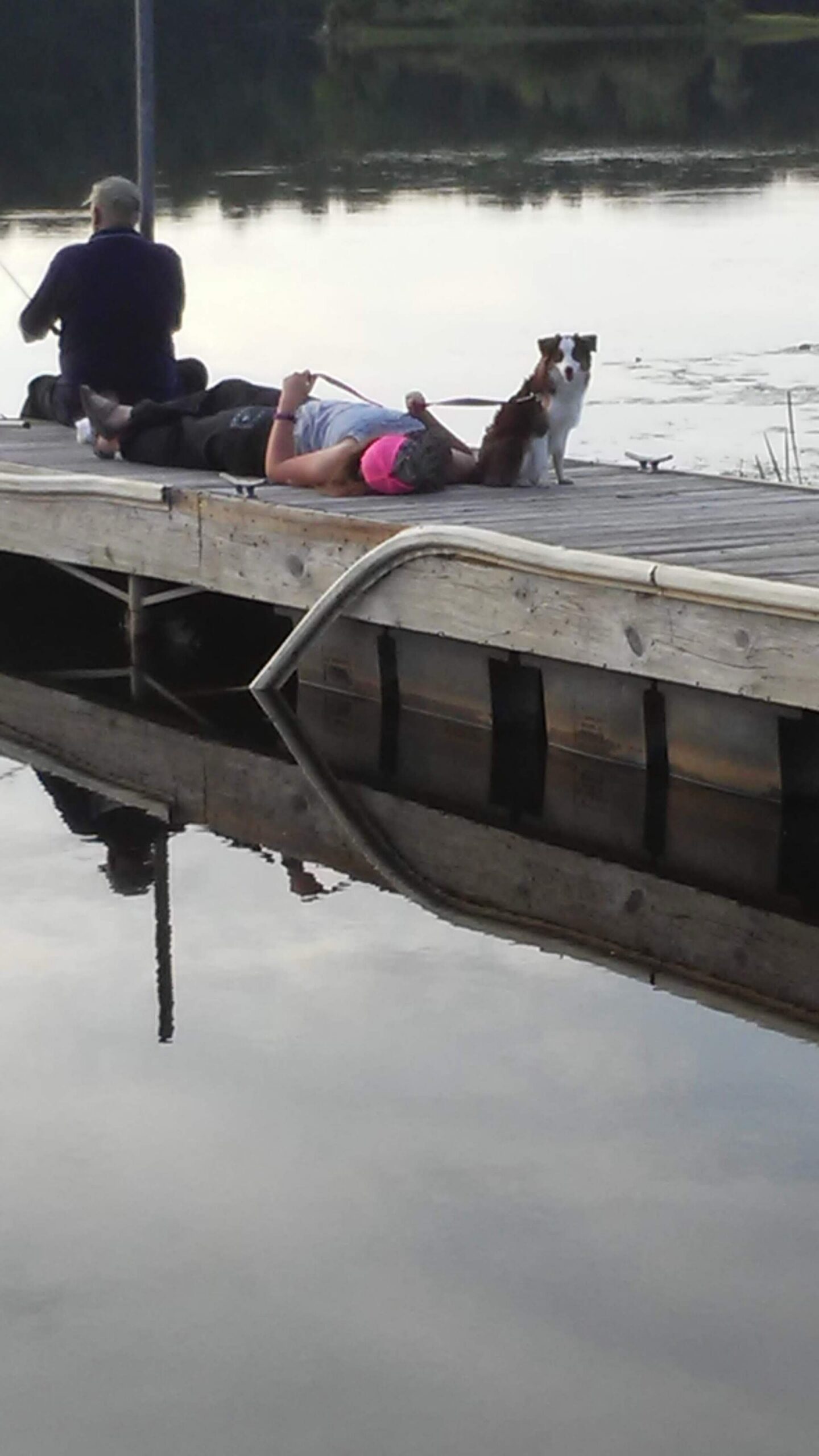 Person lying on their side on a wooden dock by a calm lake; a black-and-white dog on a leash stands nearby.