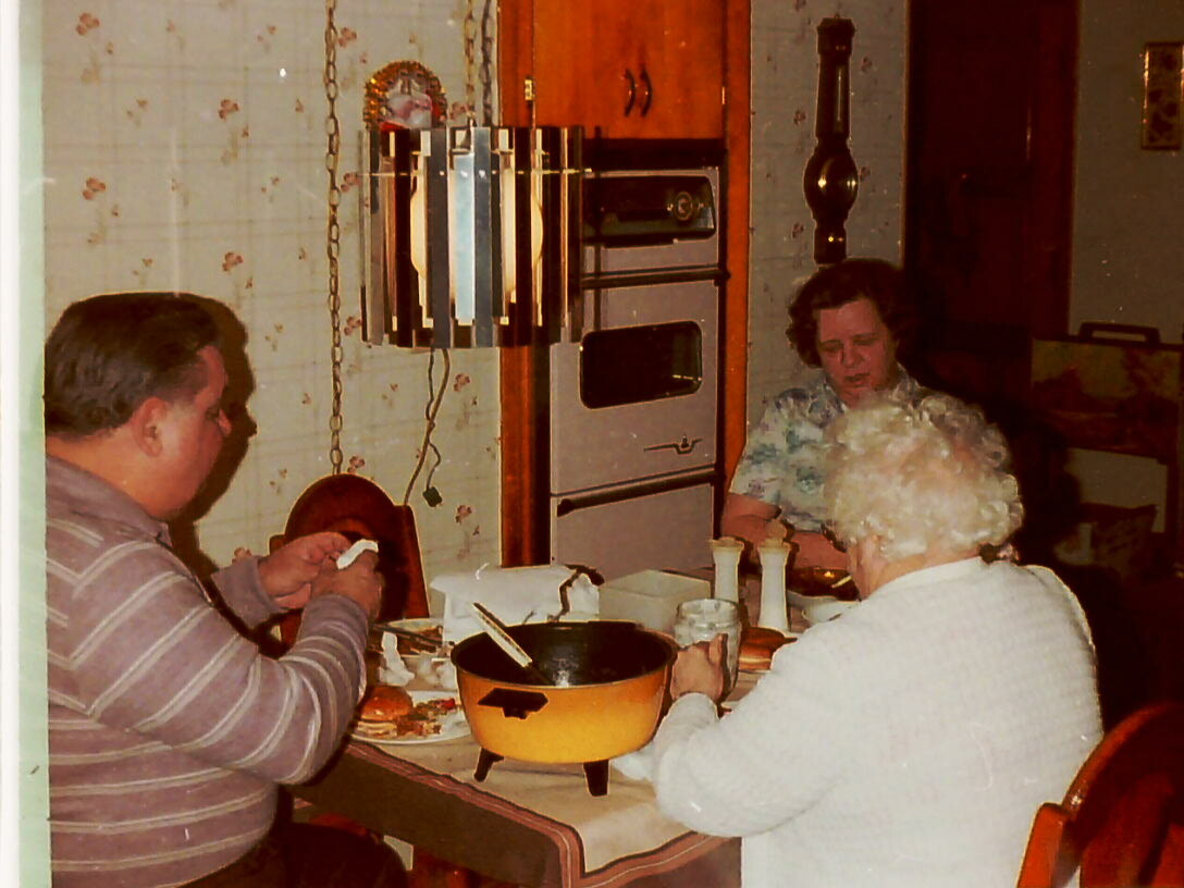 Man on the left and two elderly women dining at a table in a retro kitchen, with a yellow pot on the table in the foreground.