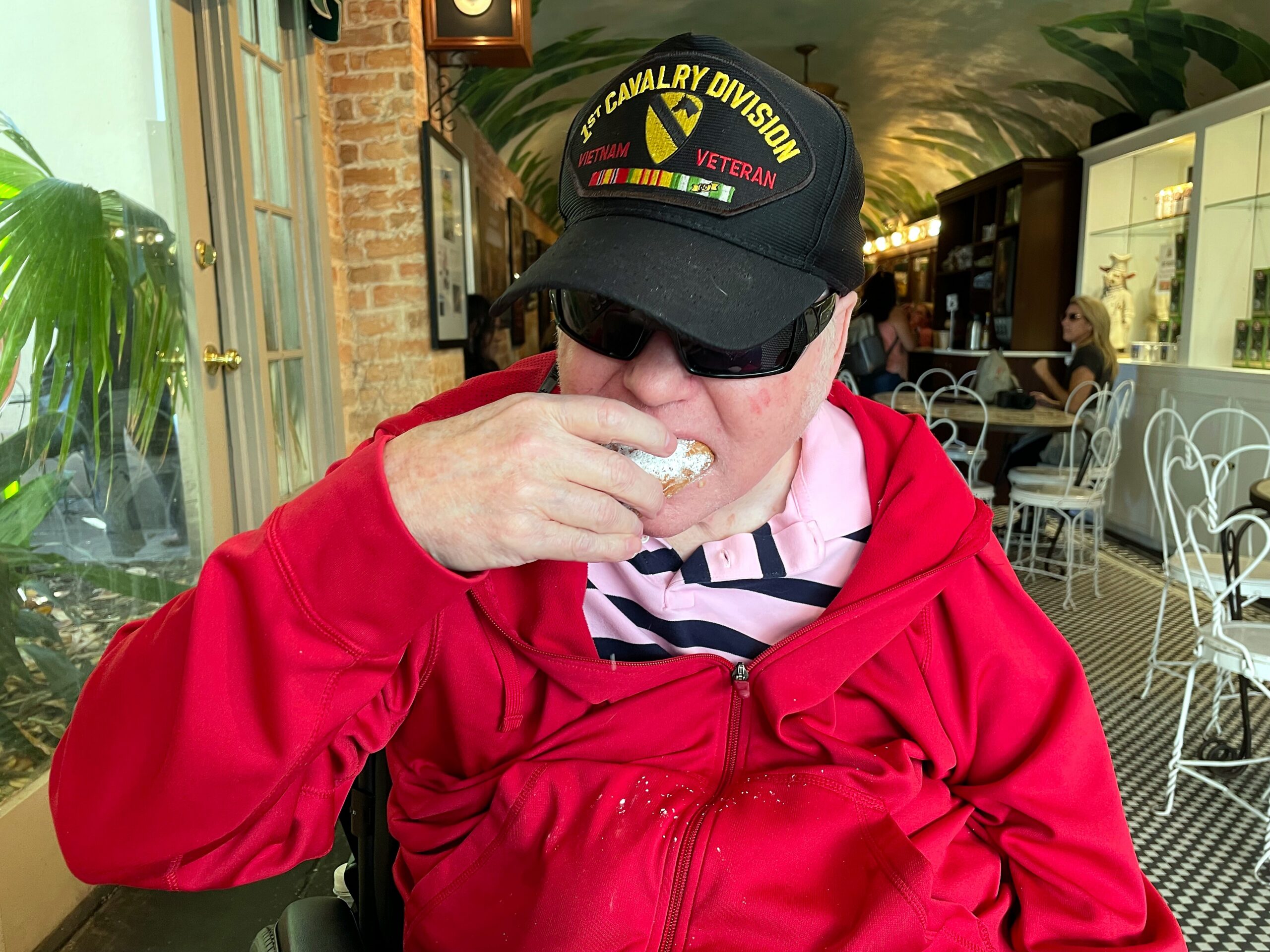 Person wearing a black cap with a Vietnam veteran patch and a red jacket eats a pastry at a cafe, with a pink striped shirt visible.