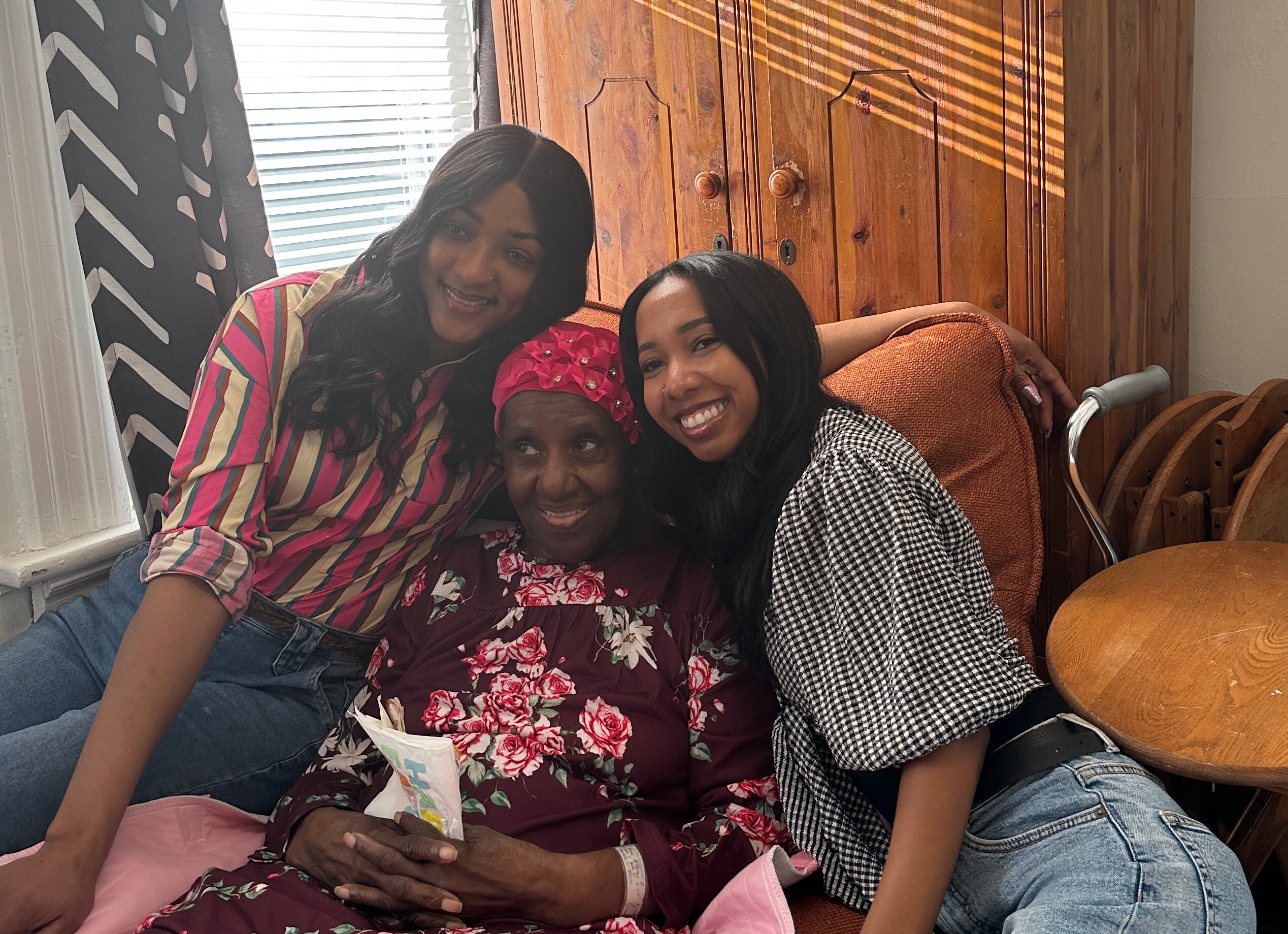 Three women sit close together on a couch indoors, smiling at the camera.