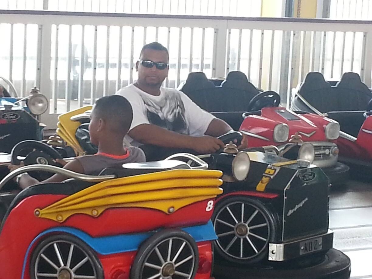 Two people ride bumper cars in an indoor amusement area, a man in sunglasses driving a black car beside a child in a red-and-blue car with yellow streams on top.