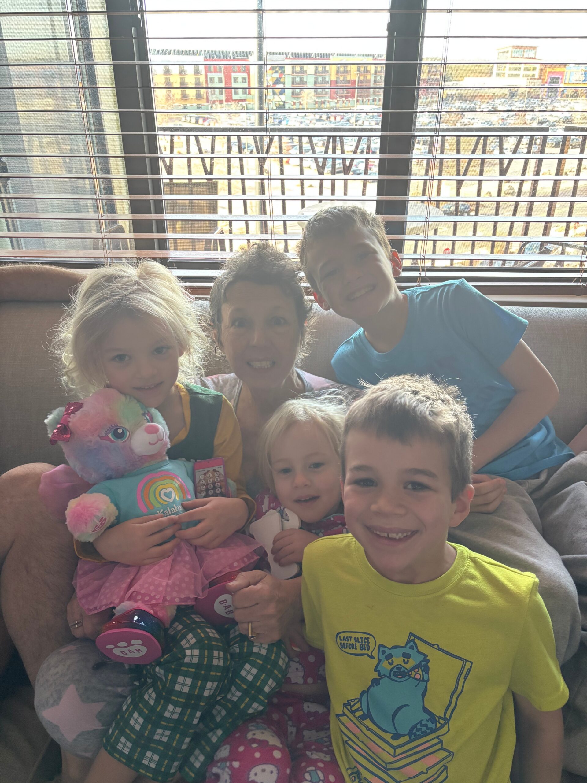Children smiling on a couch by a large window with horizontal blinds; a girl holds a colorful stuffed unicorn toy.