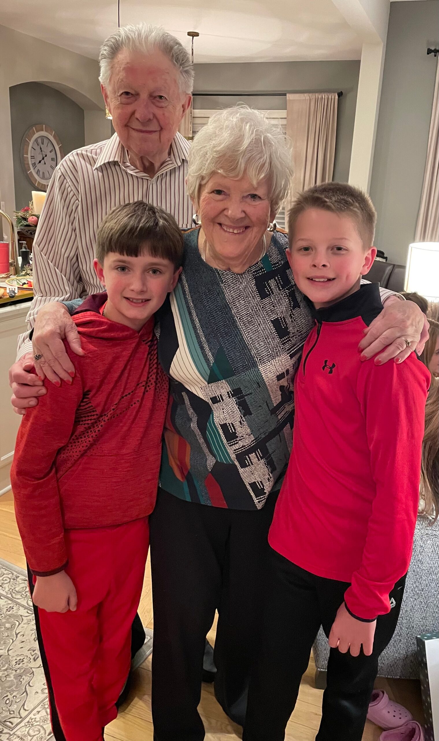An elderly woman with short white hair standing with two young boys indoors, all smiling at the camera, arms around each other in a cozy living room.