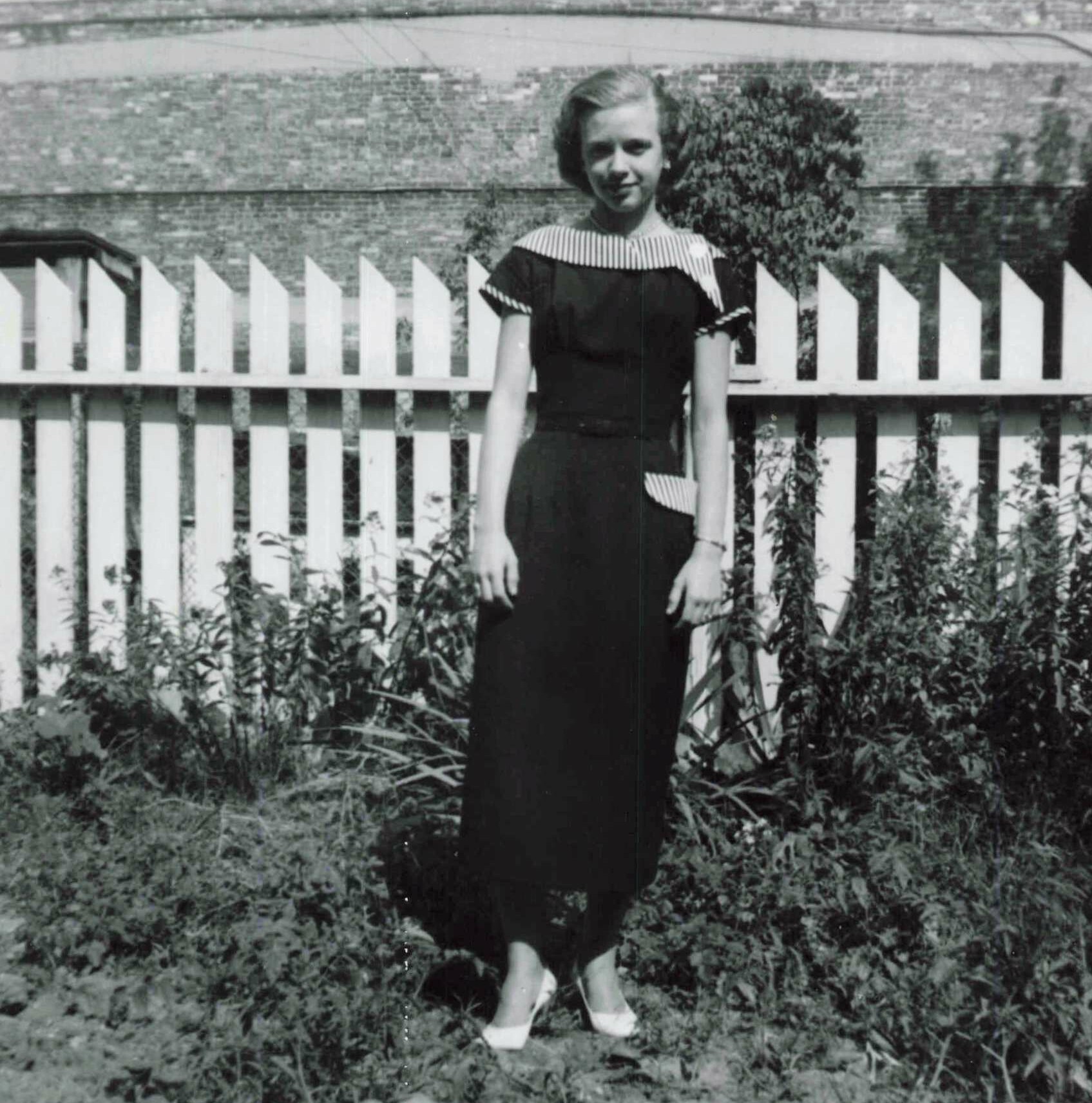 Woman in a vintage black dress with a striped collar posing outdoors in front of a white picket fence and overgrown plants.