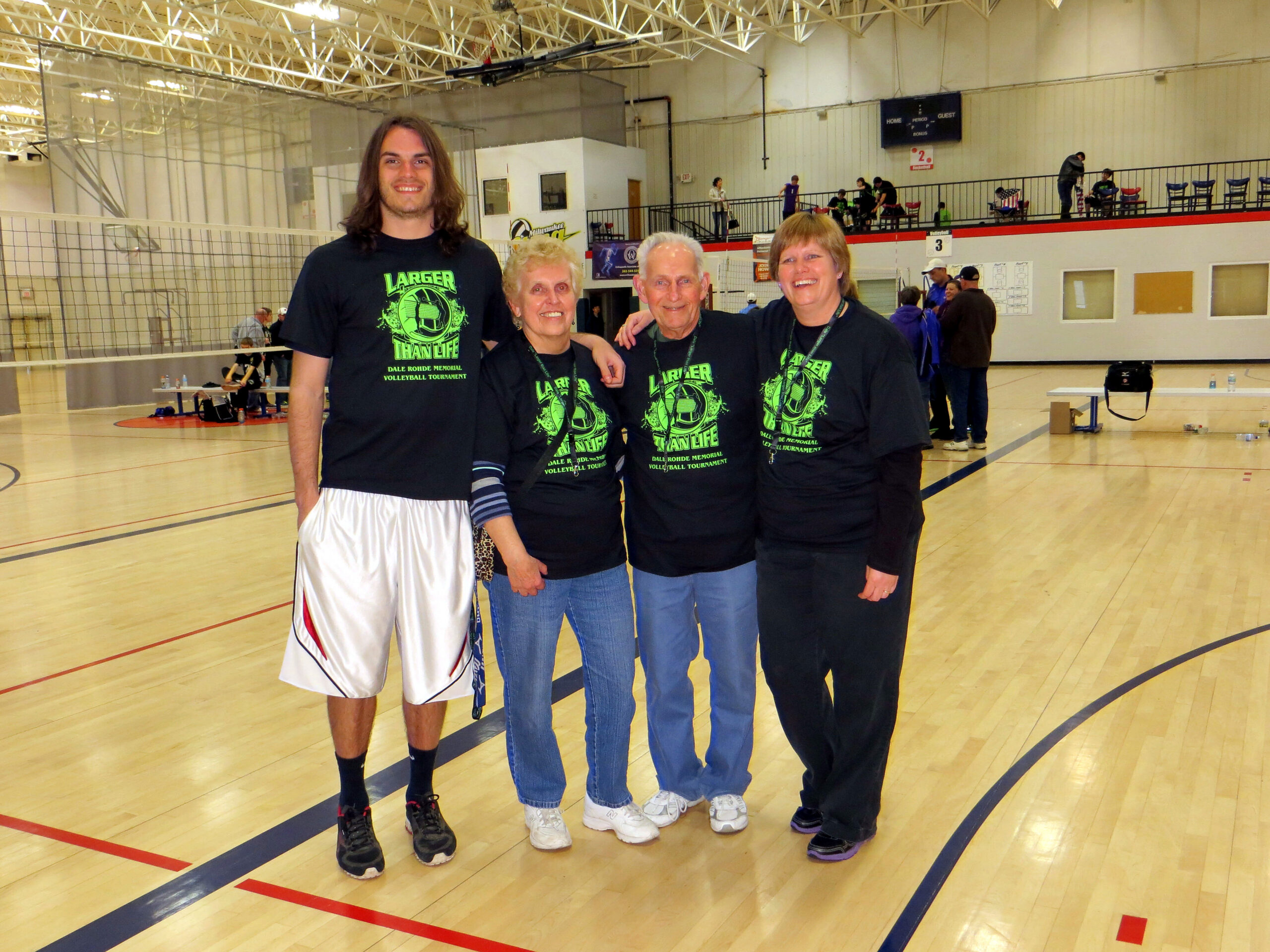 Four smiling people stand together on a gymnasium volleyball court, wearing matching black T-shirts for a tournament.