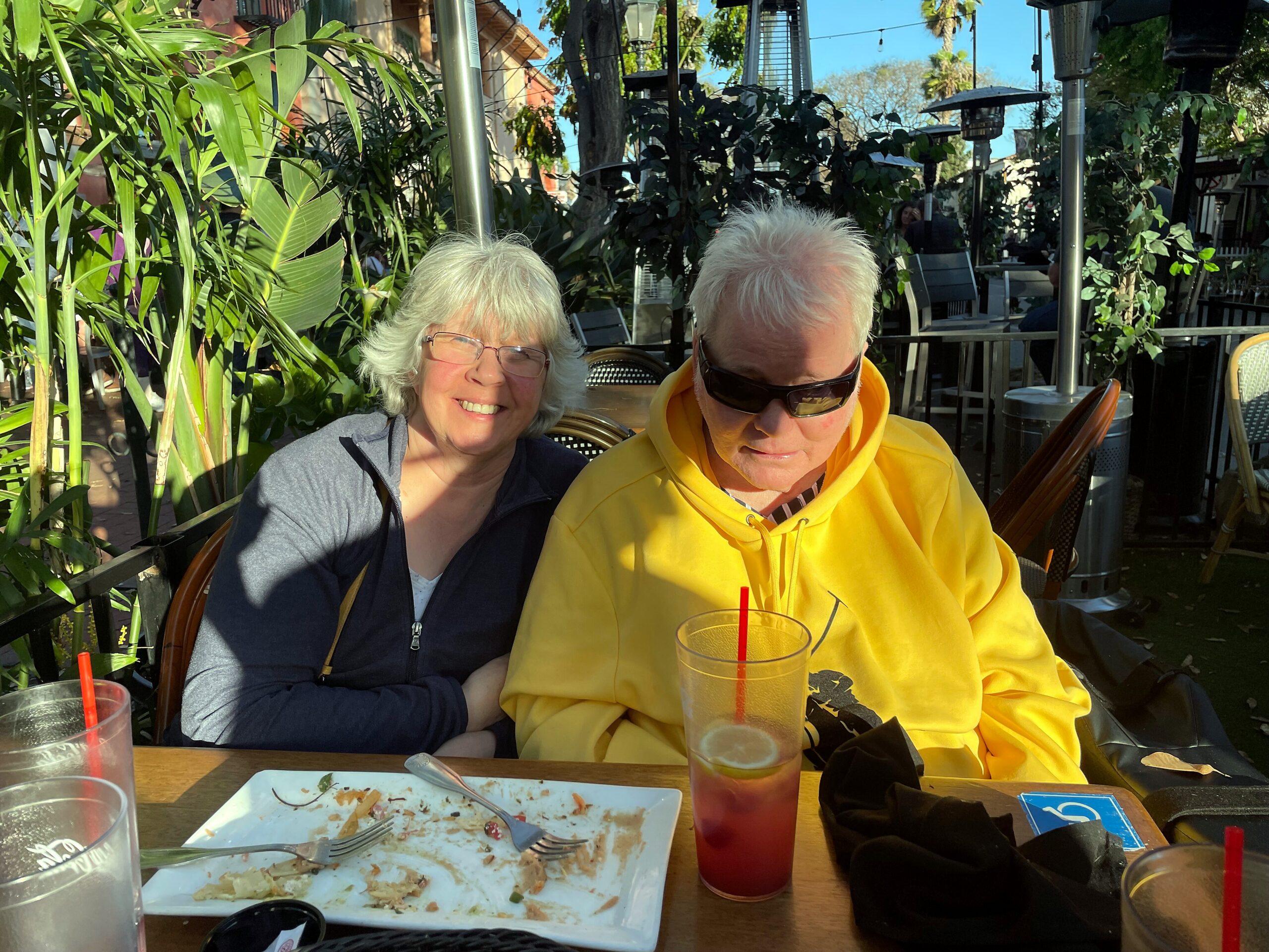 Smiling older couple sits at an outdoor cafe table with lush plants around them; woman in a gray jacket, man in a yellow hoodie wearing sunglasses ahead of a drink-filled table.