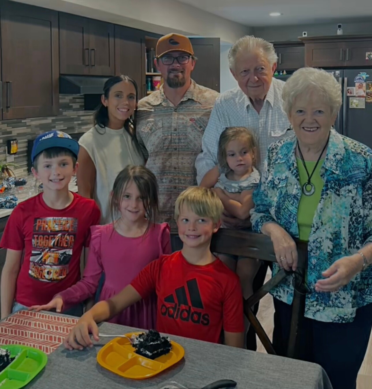 Family of eight in a kitchen posing for a group photo around a table with cake desserts visible on plates behind them on the table?