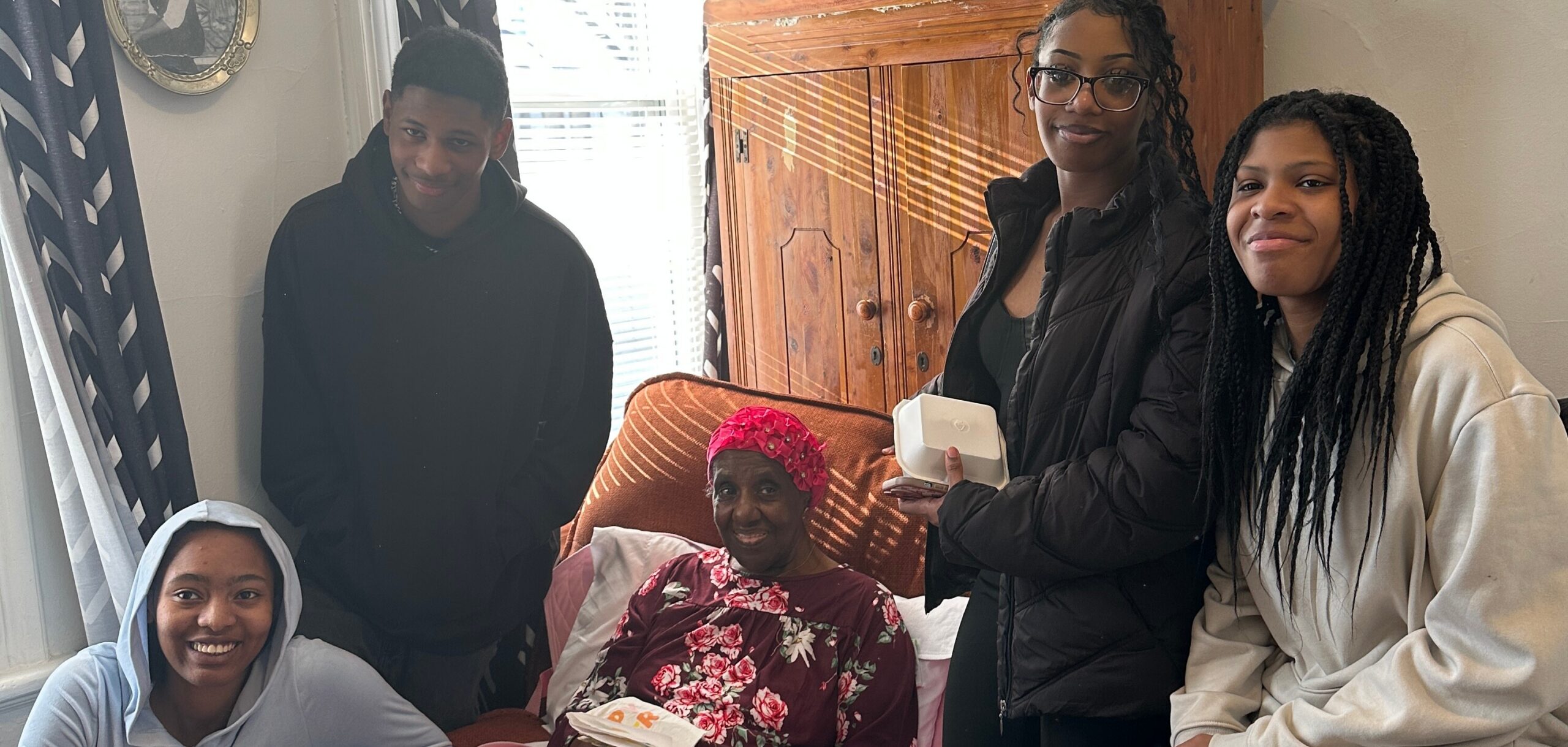 Group of five people gathered in a living room, smiling around an elderly woman sitting on an orange sofa wearing a floral blouse and red head wrap.