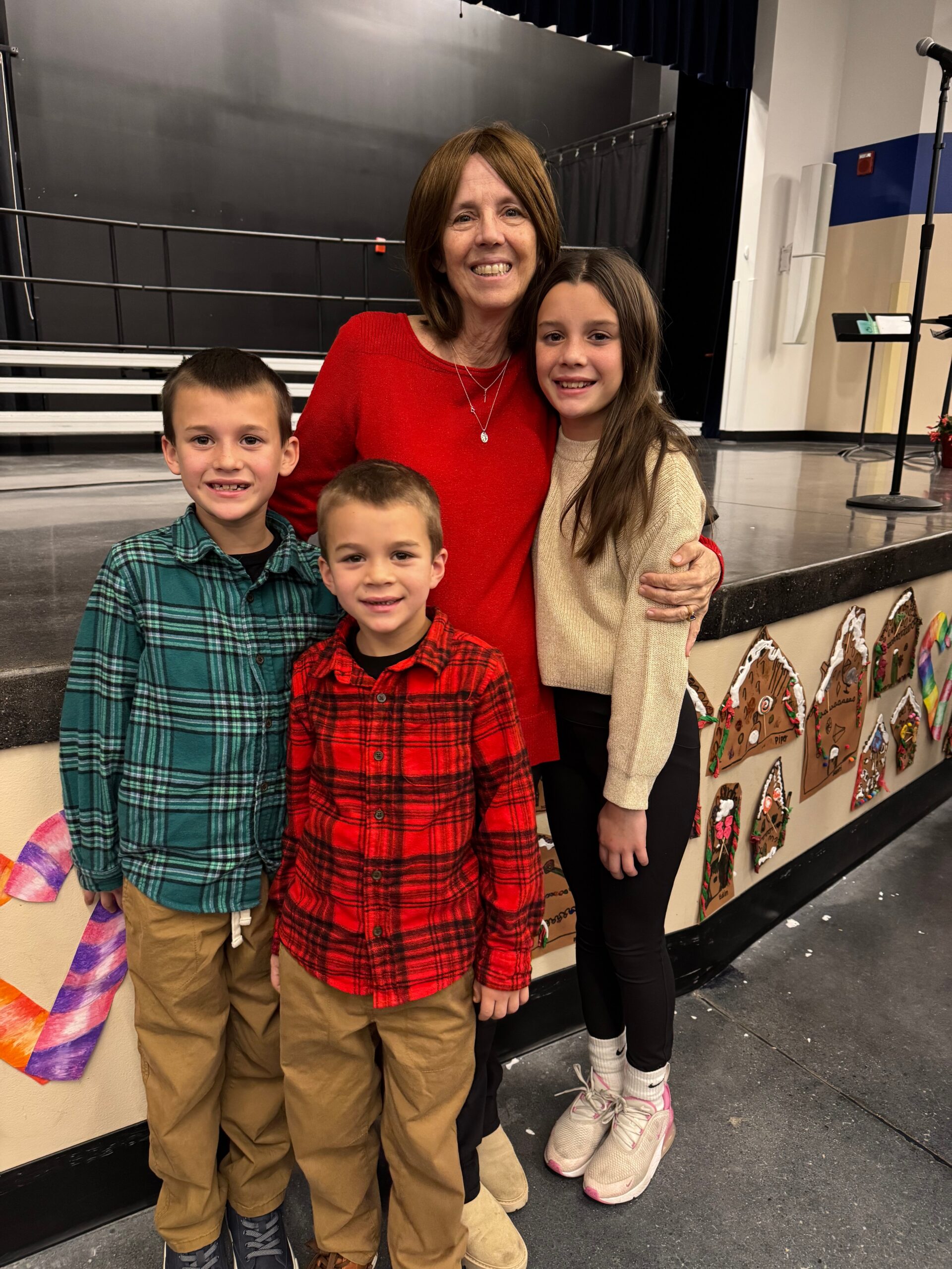 Smiling woman in a red sweater posing with three children in front of a school stage backdrop backstage at an event surface level area.