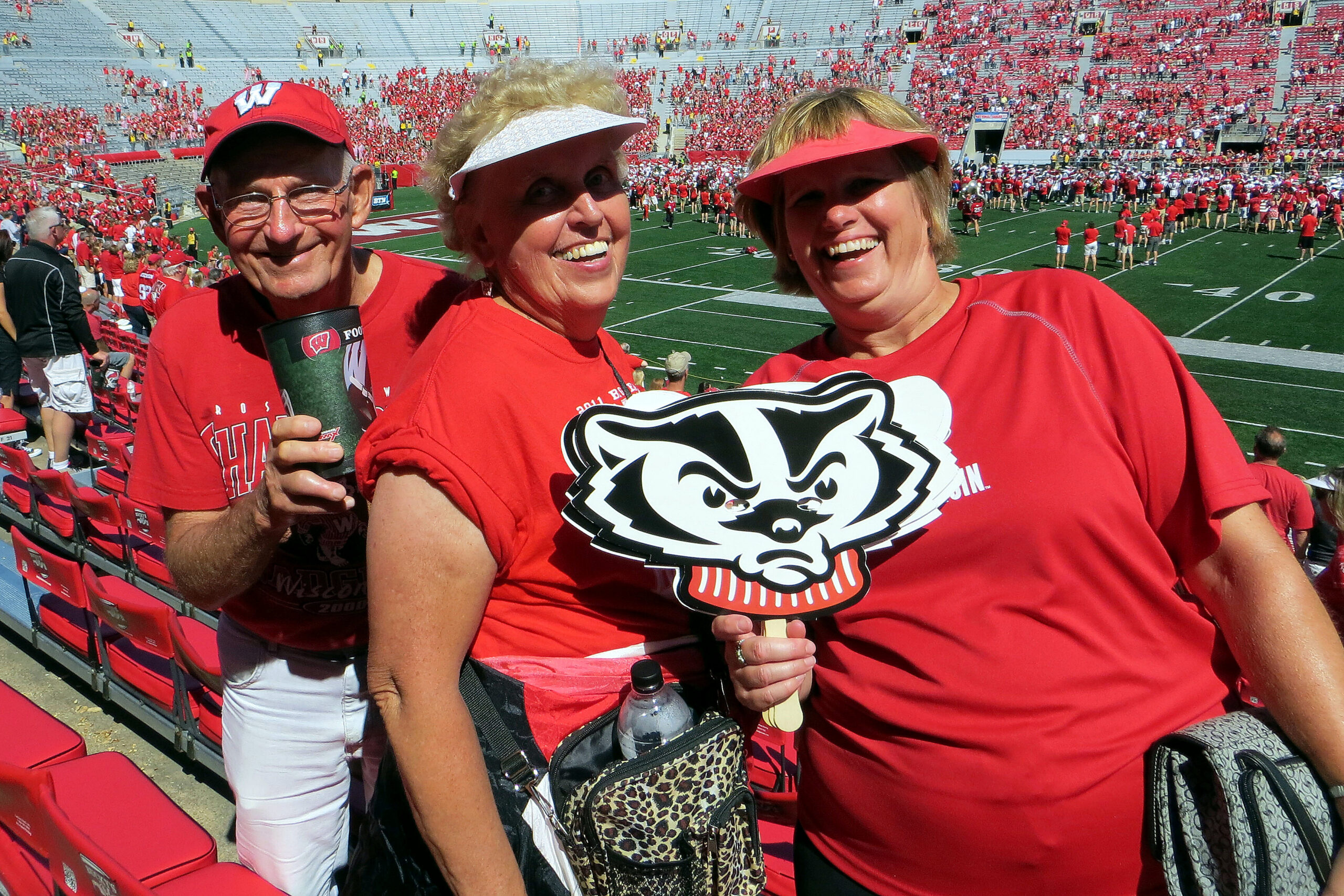 Three smiling fans in red shirts posing for a selfie at a college football stadium, one holding a large mascot cutout and a water bottle nearby?