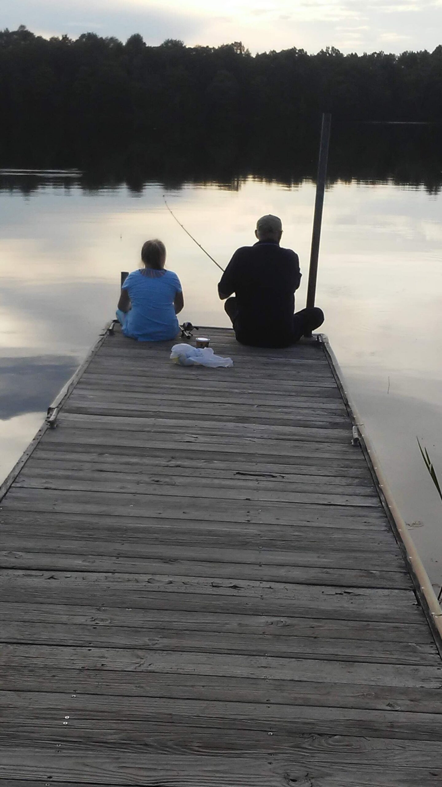 Two people sit on a weathered wooden dock by a calm lake at dusk, fishing together with a line cast into the water.