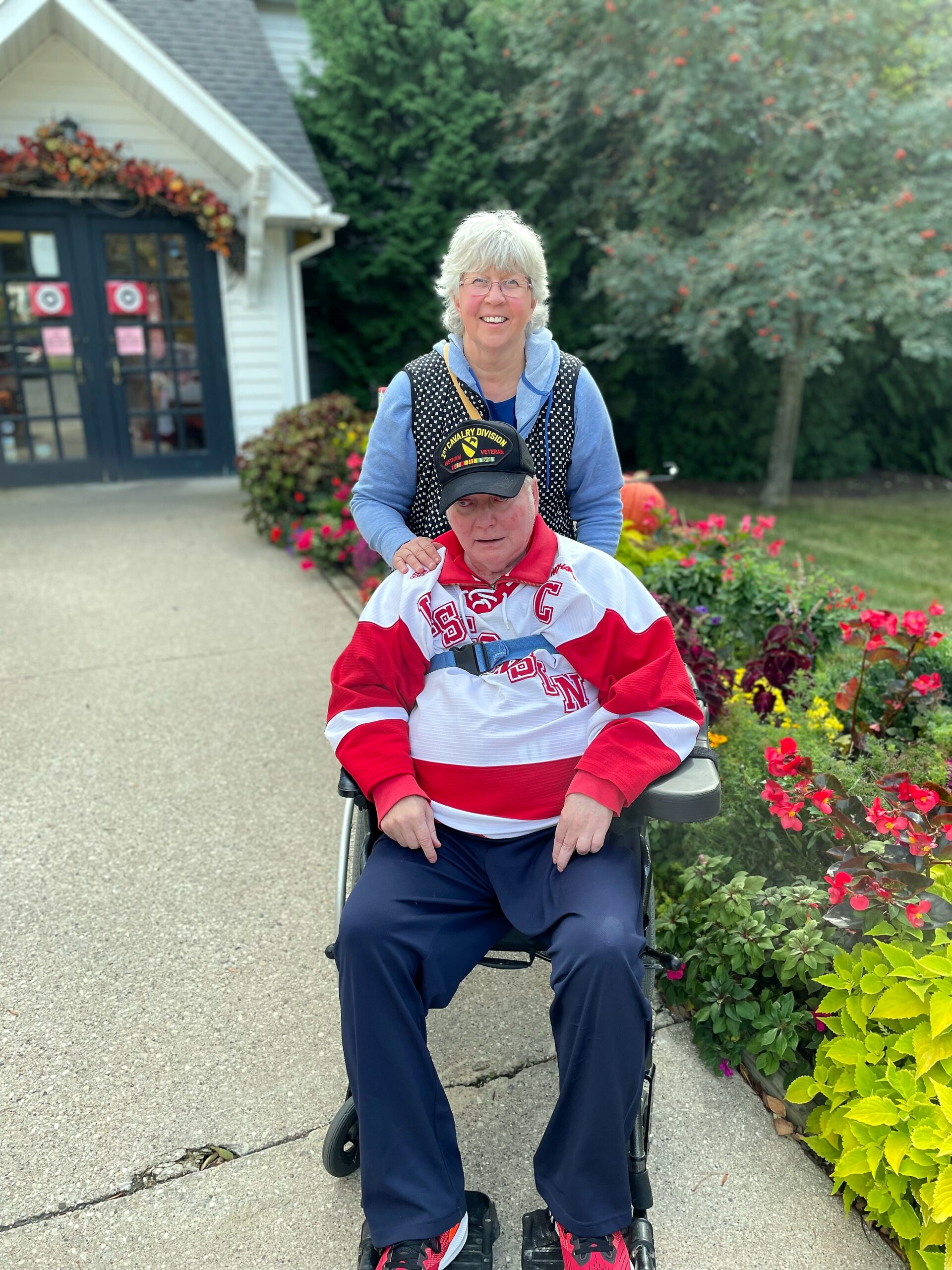 Smiling woman standing behind an elderly man in a wheelchair outdoors, beside a flower-filled garden and a building entrance.