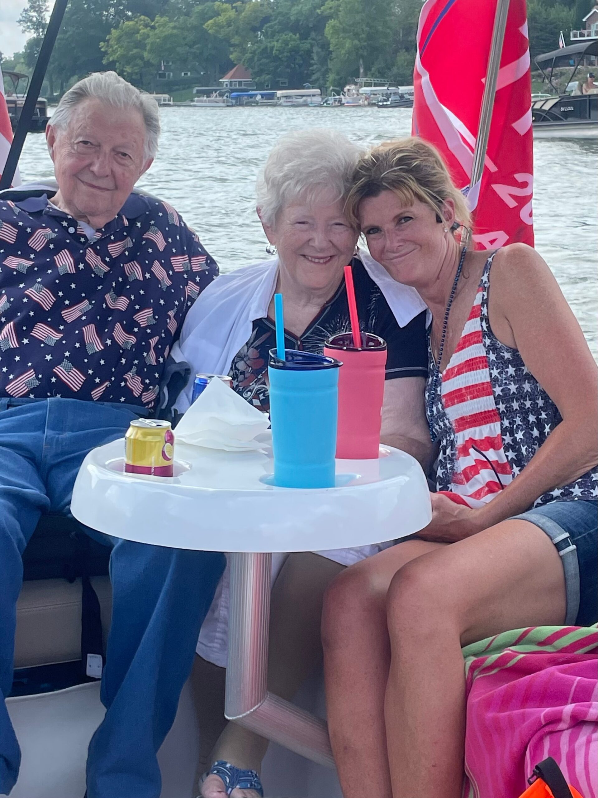 Three people on a boat by the lake, smiling for the camera; man on the left in a star-spangled shirt, two women in patriotic outfits, drinks on a white table in front.