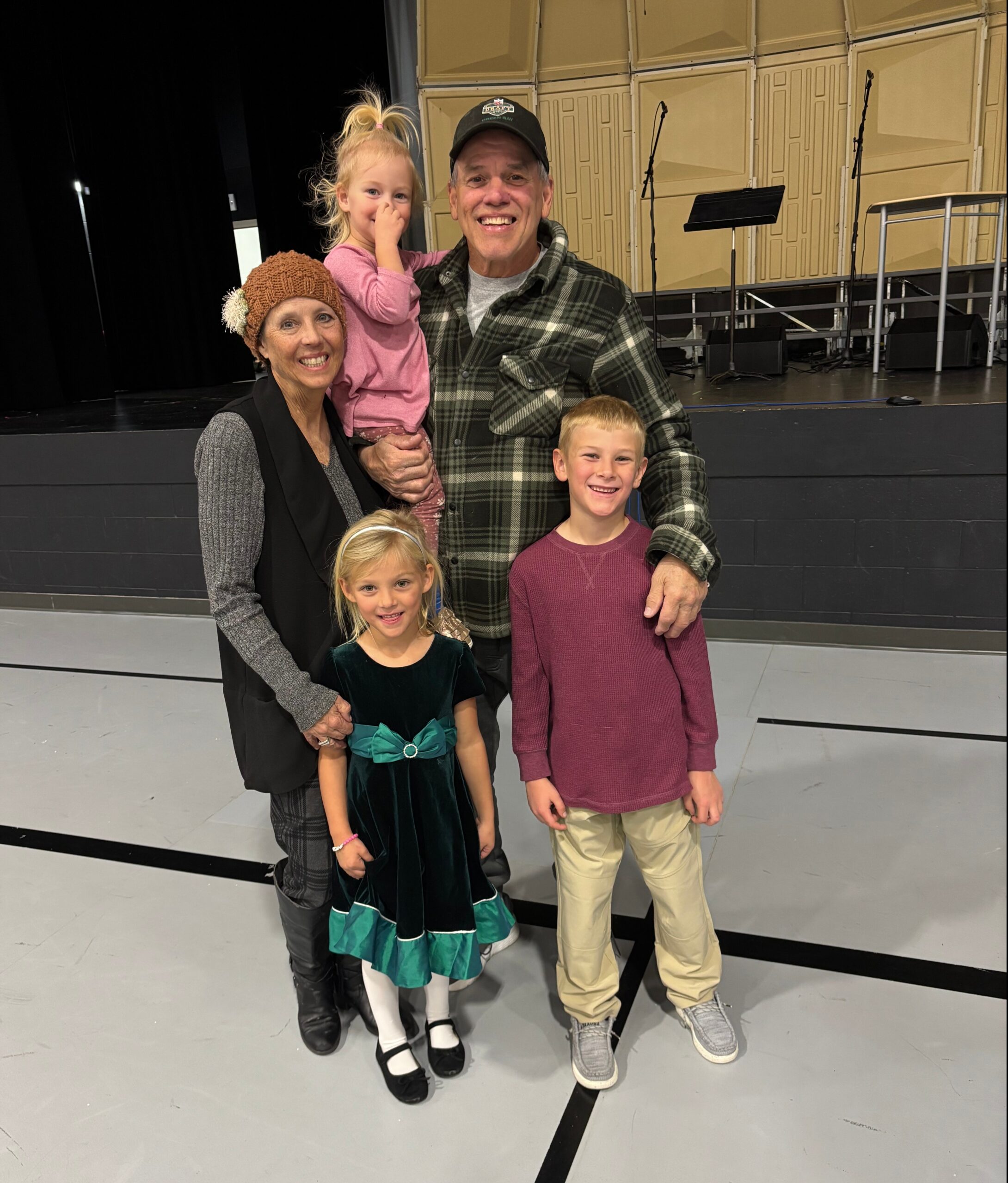 Smiling family on a stage: grandmother in a brown knit hat, a man in a plaid shirt holding a girl in pink, with two kids standing in front and to the side. Behind them are microphones and stage equipment.