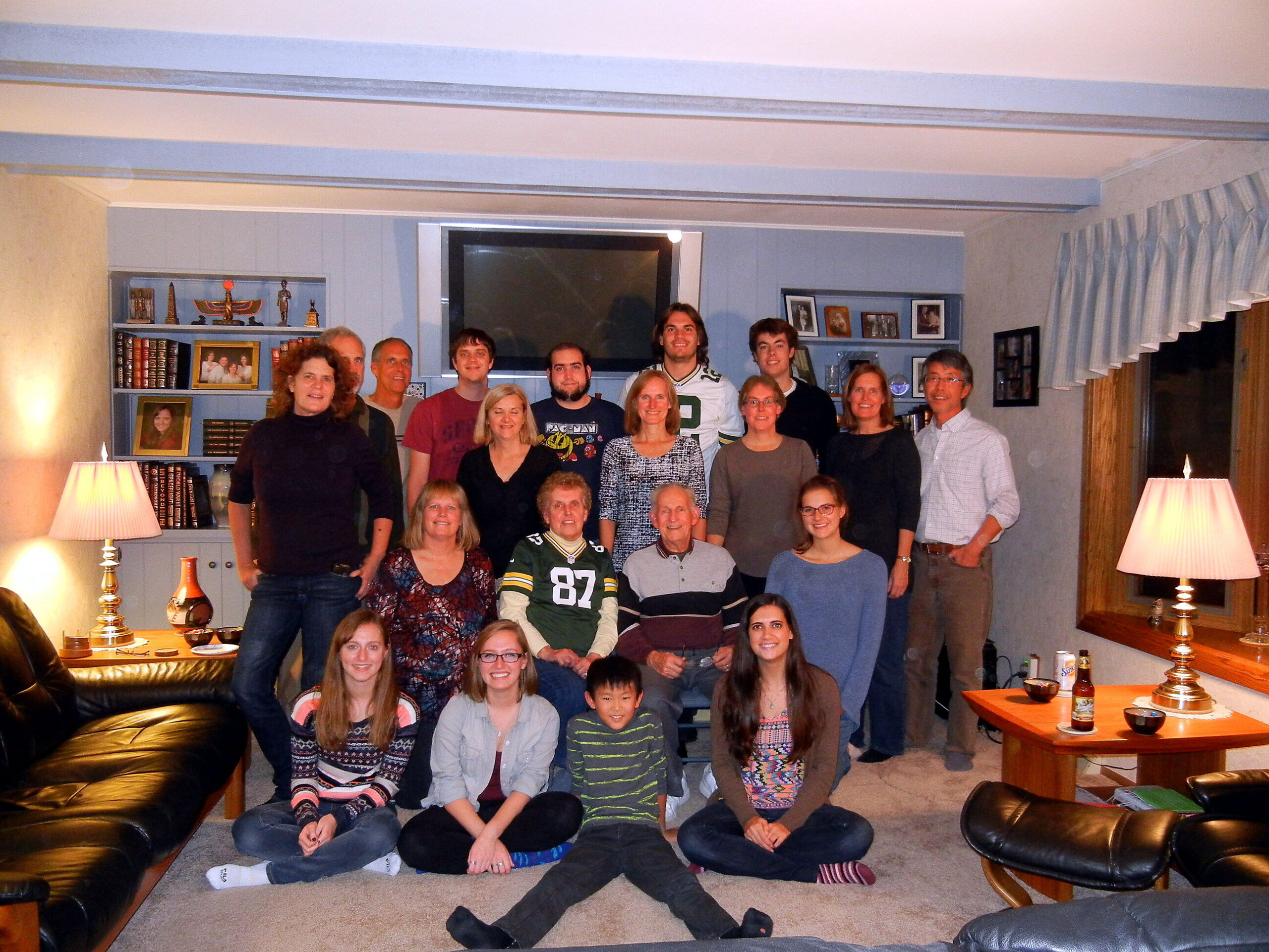 Group of about 20 people posing for a photo in a cozy living room with blue walls, shelves, and a large TV above the fireplace area.