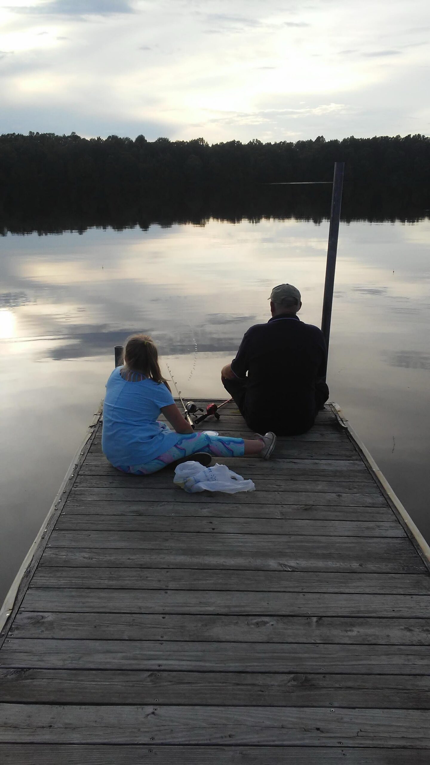 Man and girl fishing off a wooden dock on a calm lake at sunset, with trees in the distance and cloudy sky reflected on the water.
