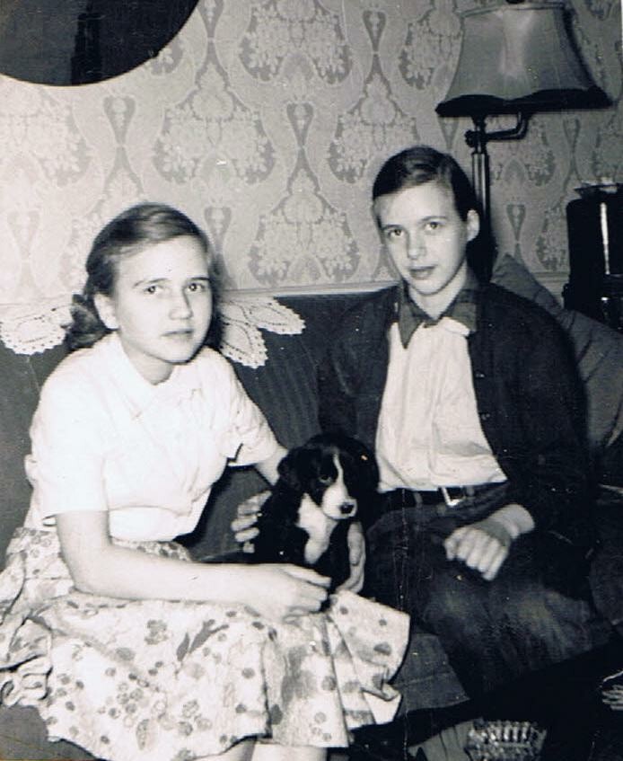Two teenagers sit on a couch with a small black‑and‑white puppy between them in a vintage living room.