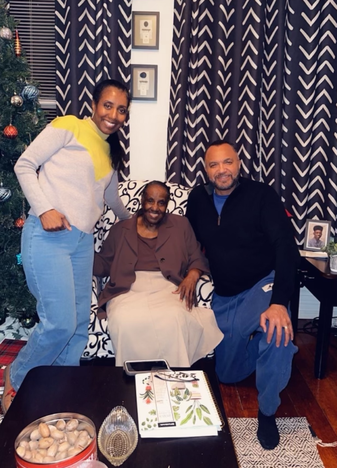 Three adults posing with an elderly woman in a living room, standing on either side of her near a patterned chair and Christmas tree.