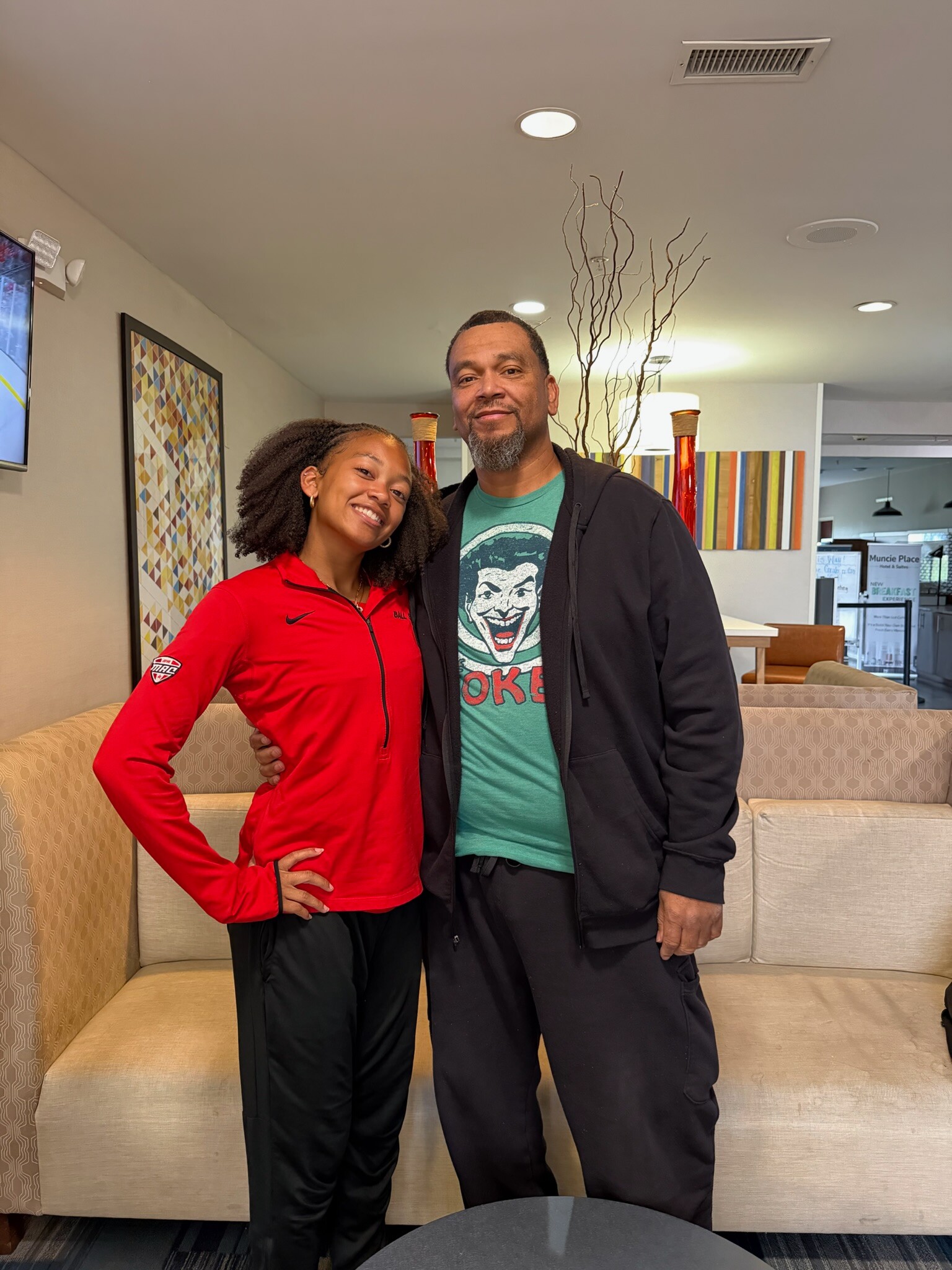 Smiling woman in a red jacket and a man in a teal Joker shirt pose together in a contemporary lounge area with beige couches and artwork behind them.