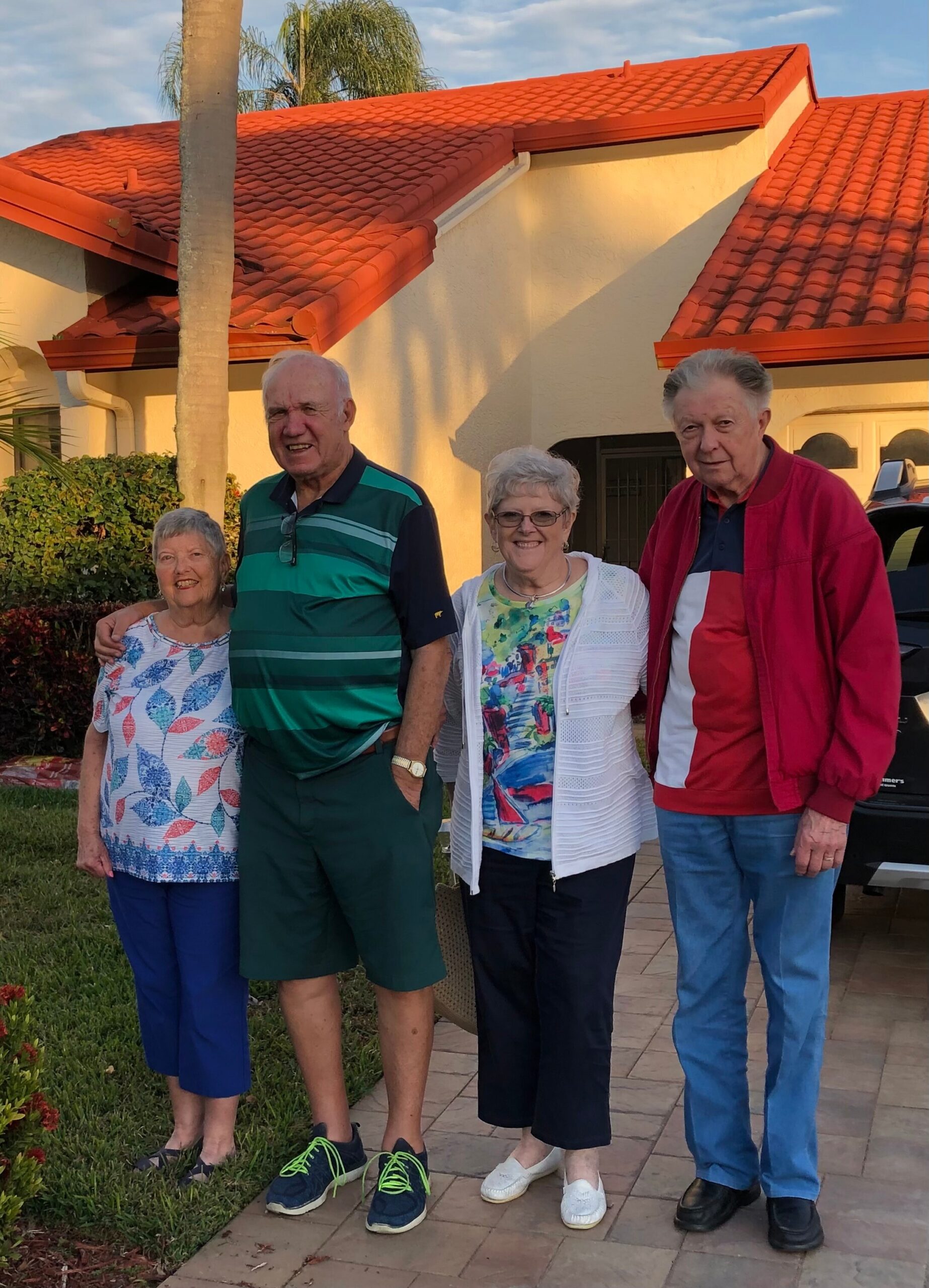Four seniors posing together outside a tan house with a red-tiled roof, smiling on a sunny day after sunset light.