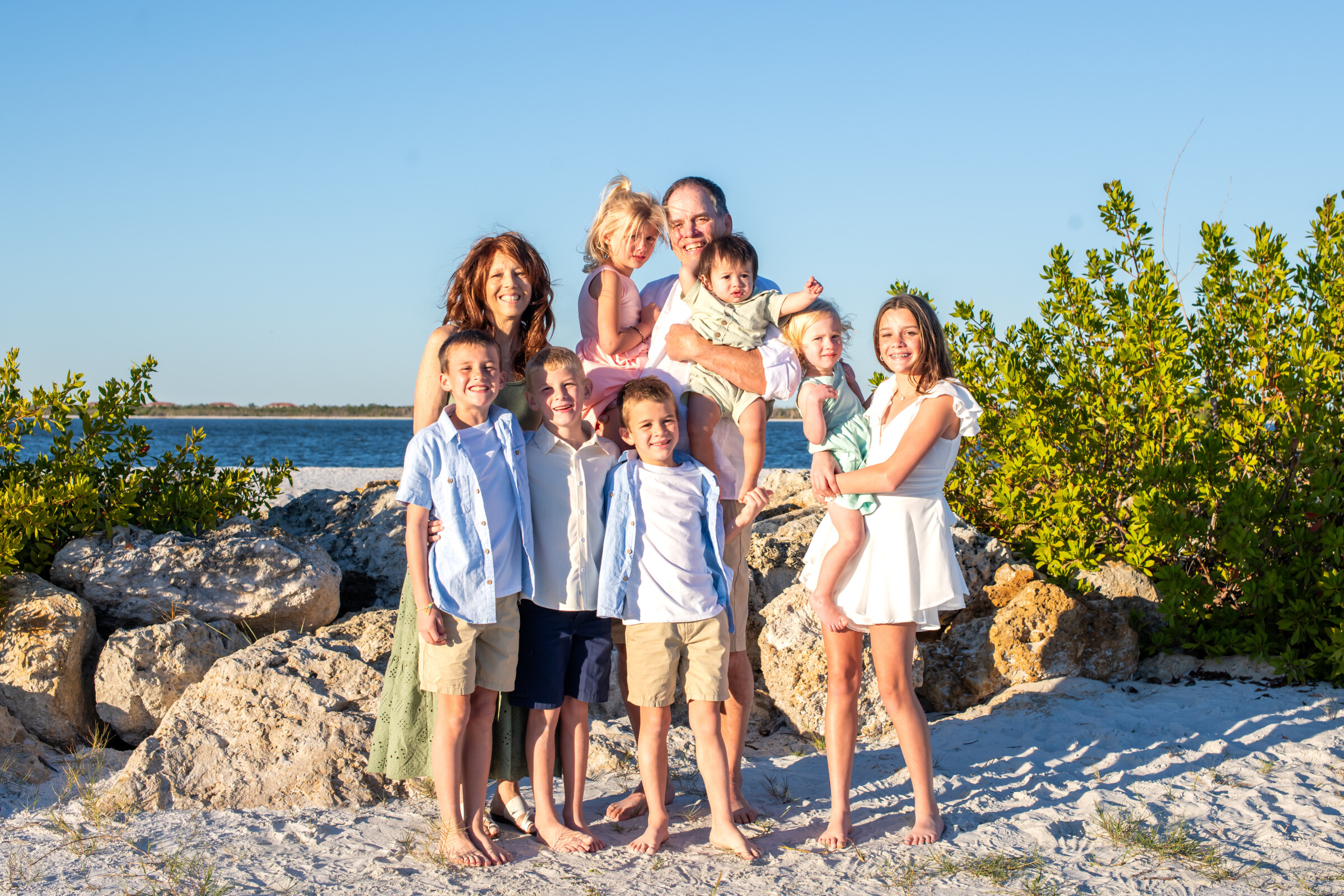 A large family group posing together on a rocky beach, smiling at the camera with the ocean and bright blue sky in the background.