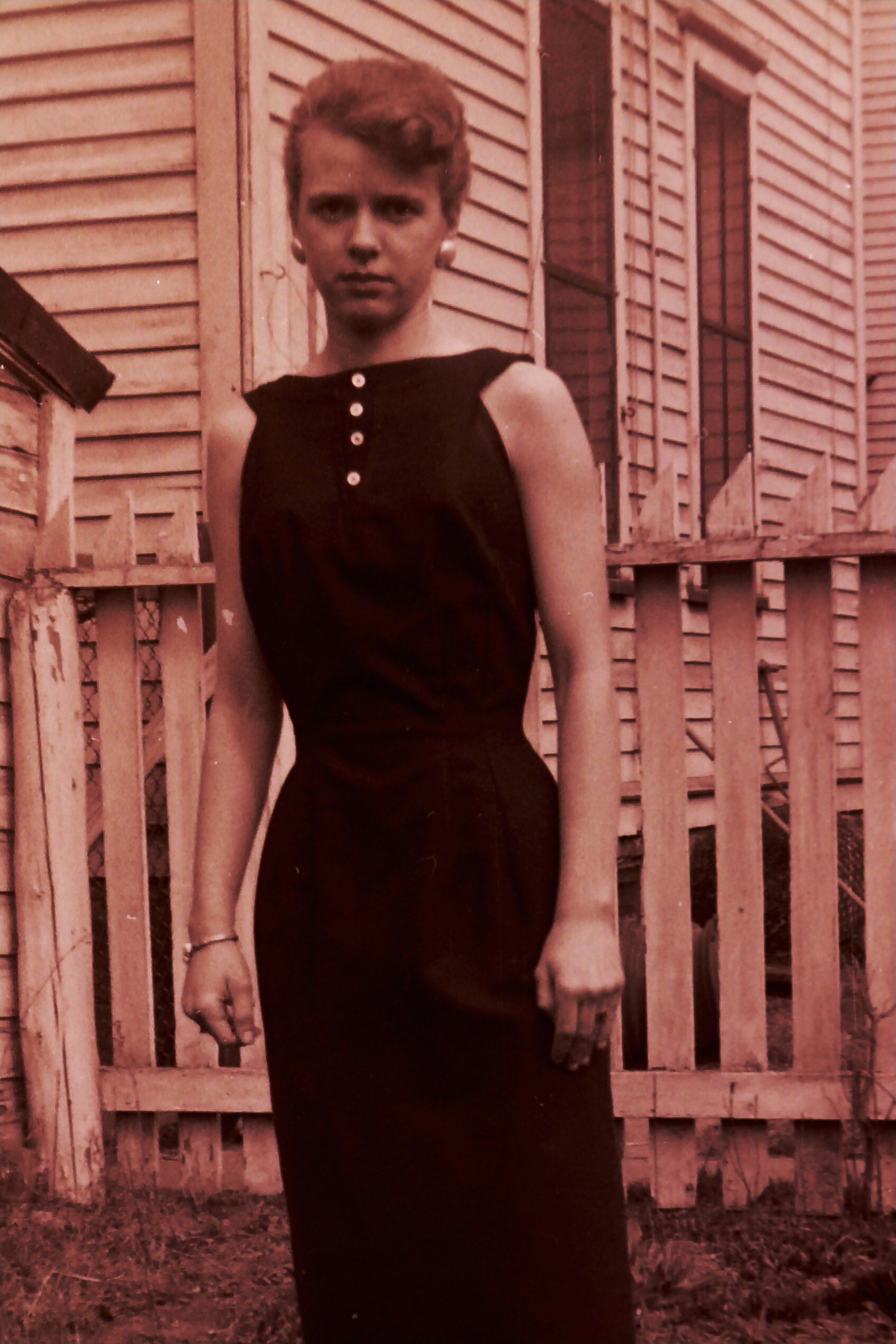 Young woman in a sleeveless dark dress with buttons, standing in front of a wooden fence and a house.