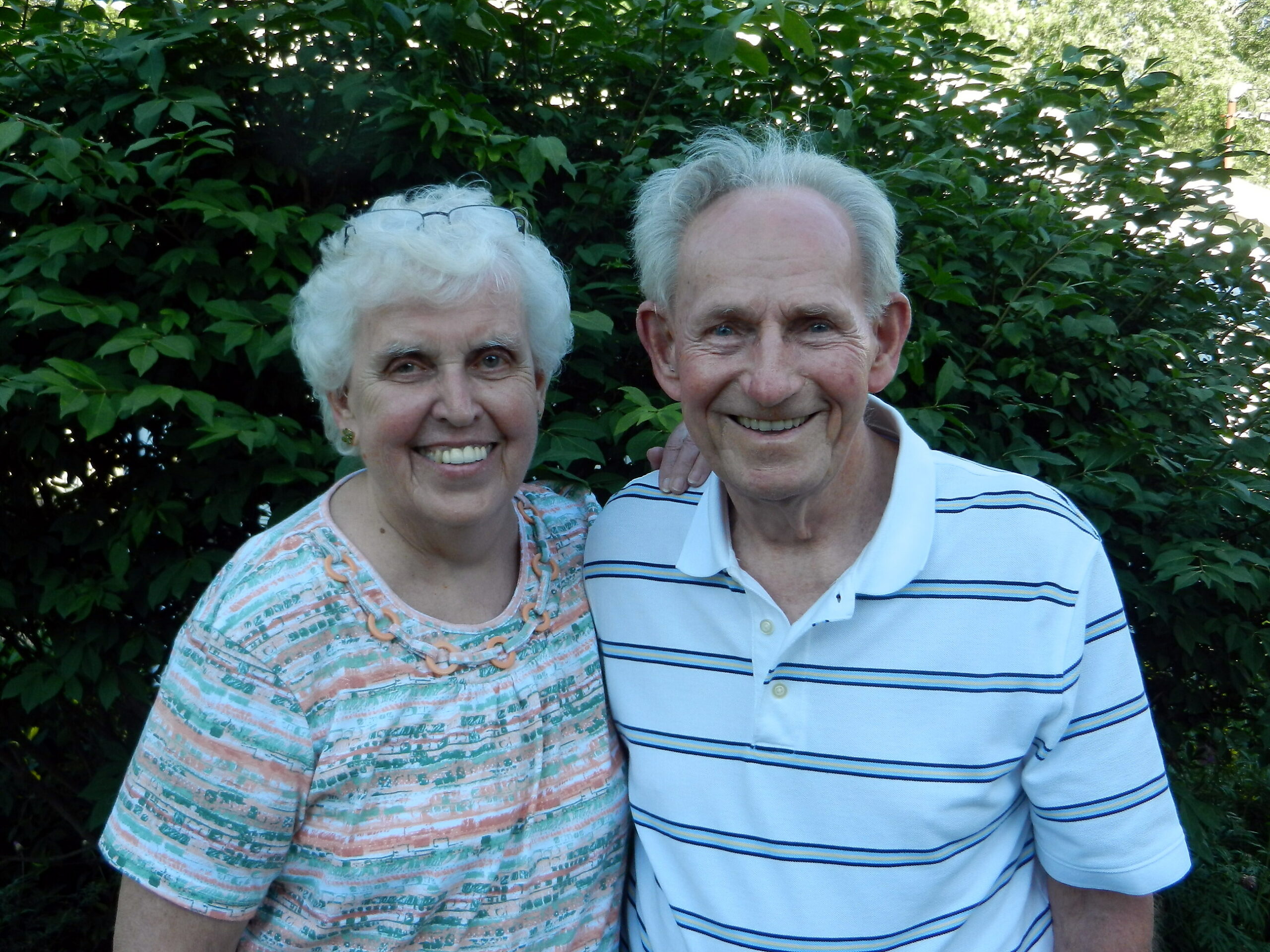 Smiling elderly couple posing outdoors in front of dense green bushes