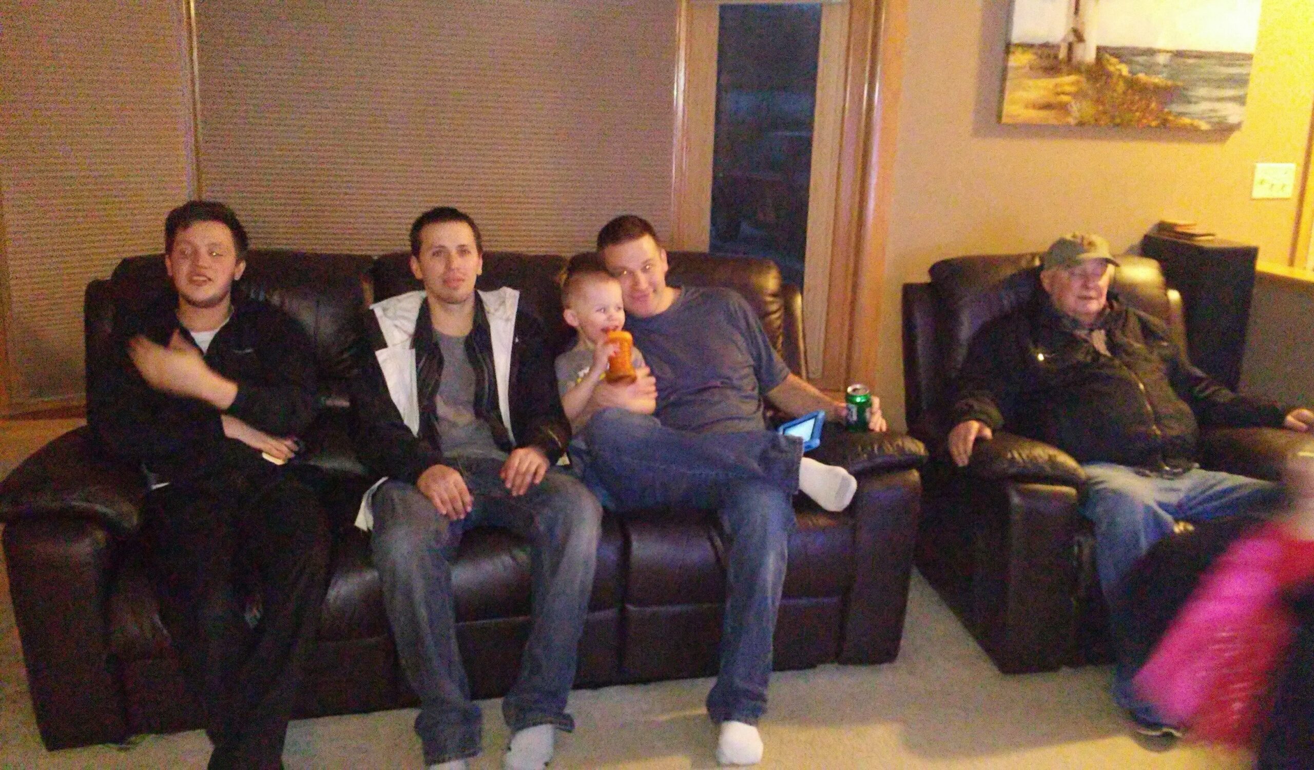 Group of five people (four adults, one child) sitting on a dark leather sofa in a living room, some holding drinks and chatting while an older man sits to the right.