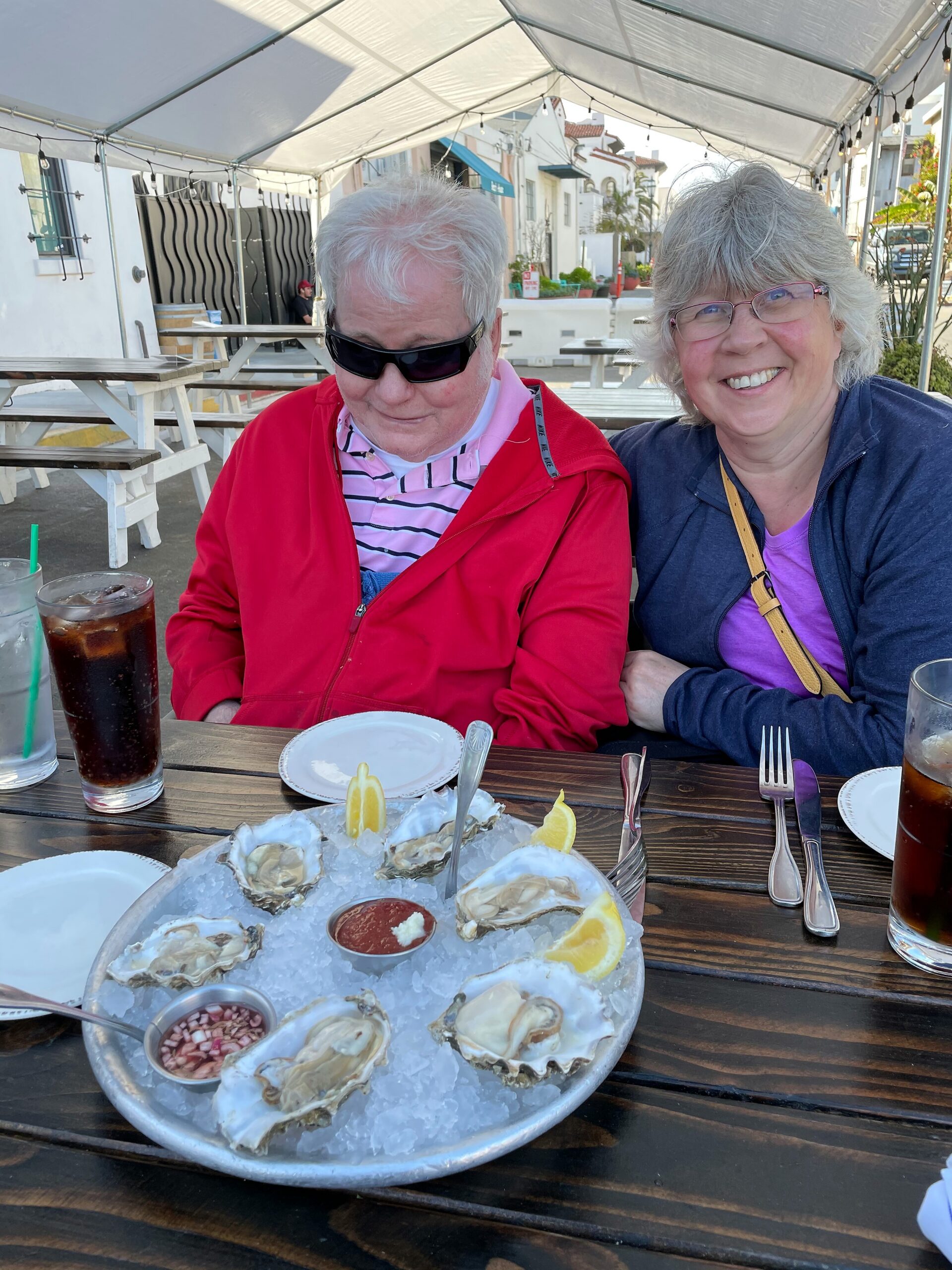 Two smiling adults sit at a wooden table under a covered outdoor dining area, with a large tray of oysters on ice in front of them.