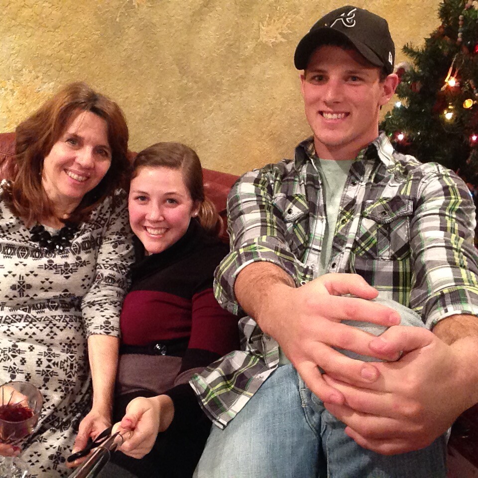 Three people sit on a couch smiling for a photo, with a Christmas tree visible to the right in a cozy room.